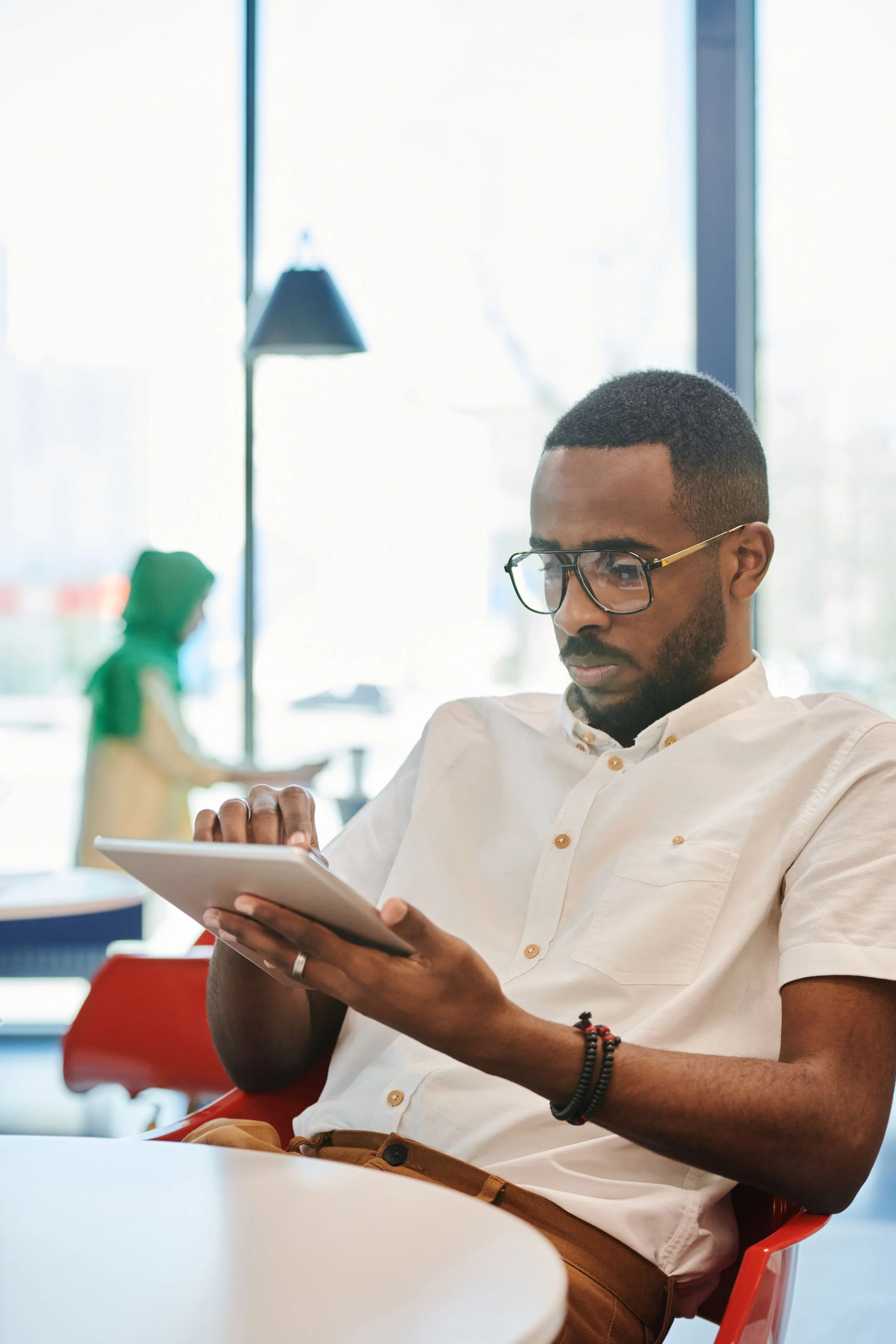 A man with short curly hair and glasses wearing a white shirt sitting in a bright room using a tablet, with a woman in a green hoodie in the background.