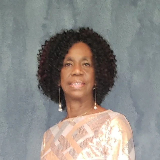 A woman with curly hair wearing a peach-colored dress with a floral pattern, standing in front of a plain blue-gray wall.
