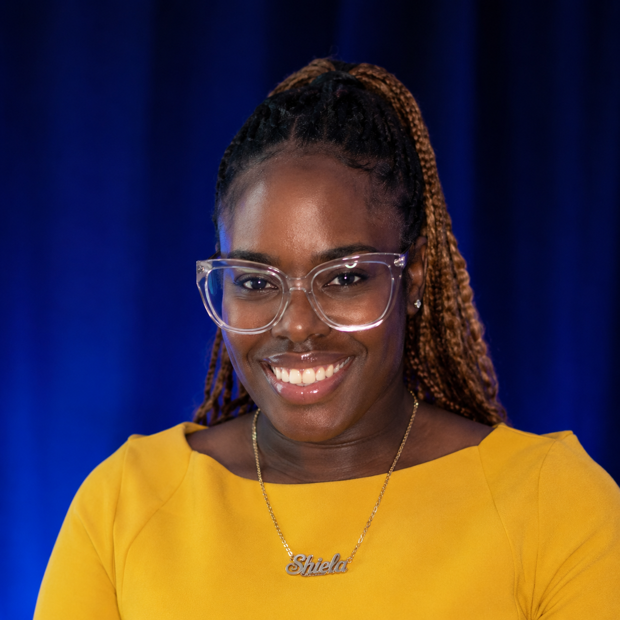 Smiling woman with glasses and braided hair, wearing a yellow top and a necklace with the name Shielda, against a dark blue background.