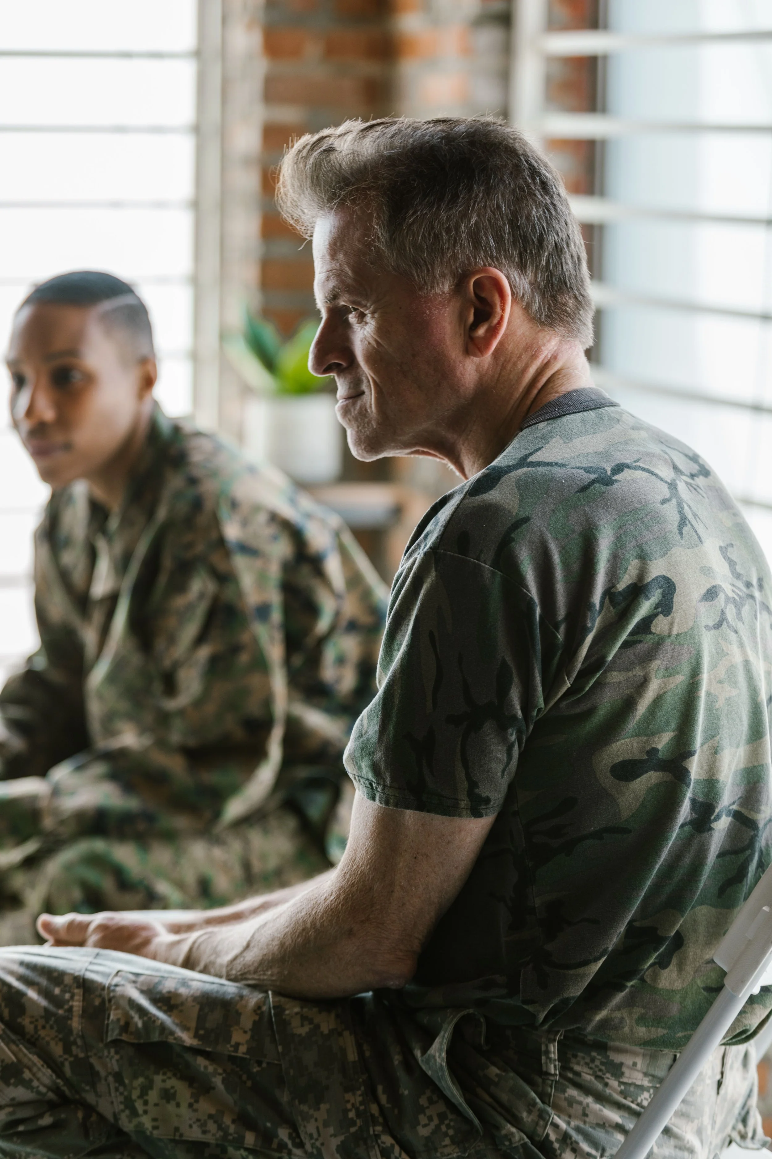 A man and a woman in military camouflage clothing sitting indoors, with a brick wall and window blinds in the background. The man is in profile with his eyes closed, and the woman is blurred in the background.