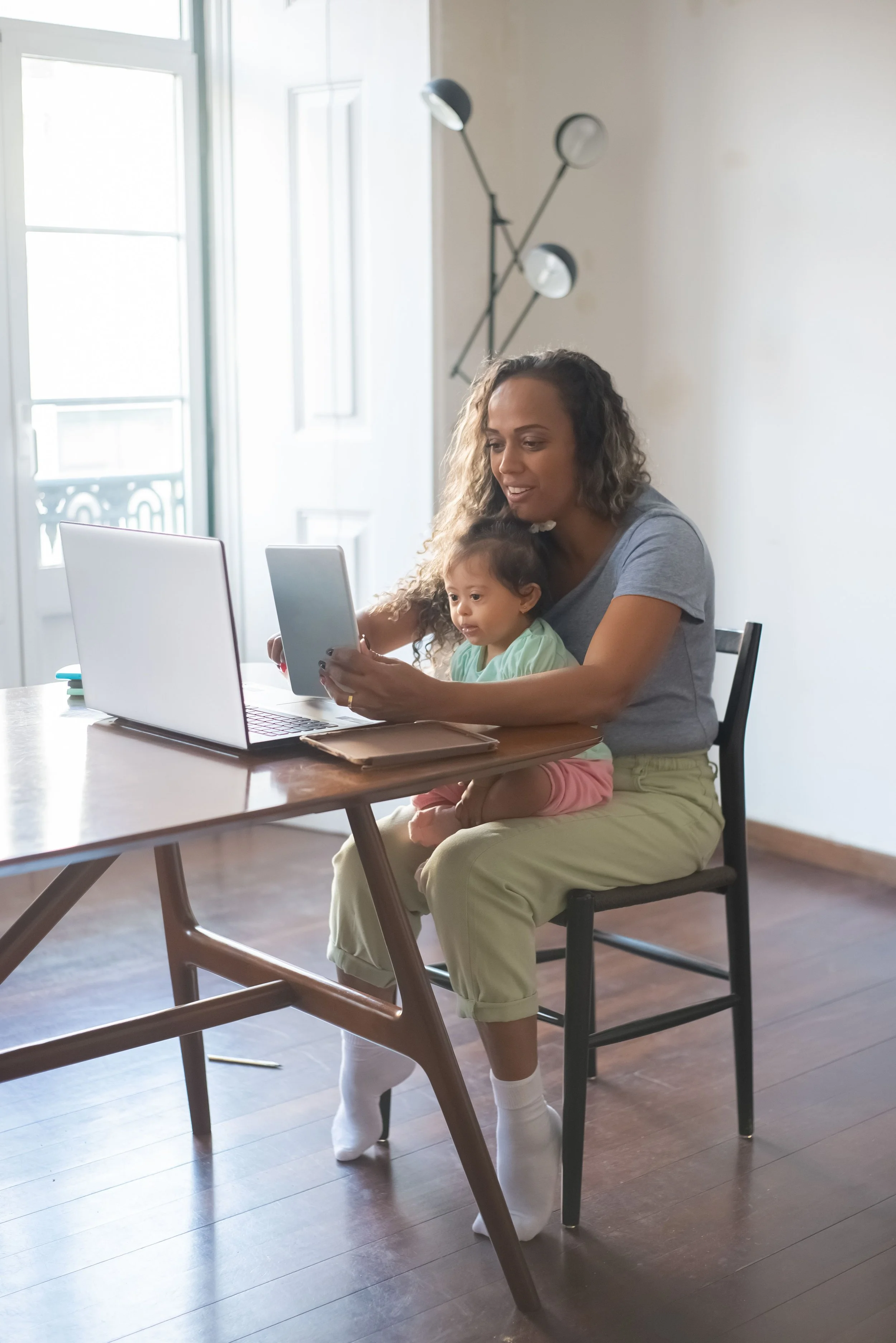 A woman sitting at a dining table with a young girl on her lap, both looking at a laptop screen. The woman is holding a tablet. They are in a bright room with large windows and a modern light fixture on the wall.