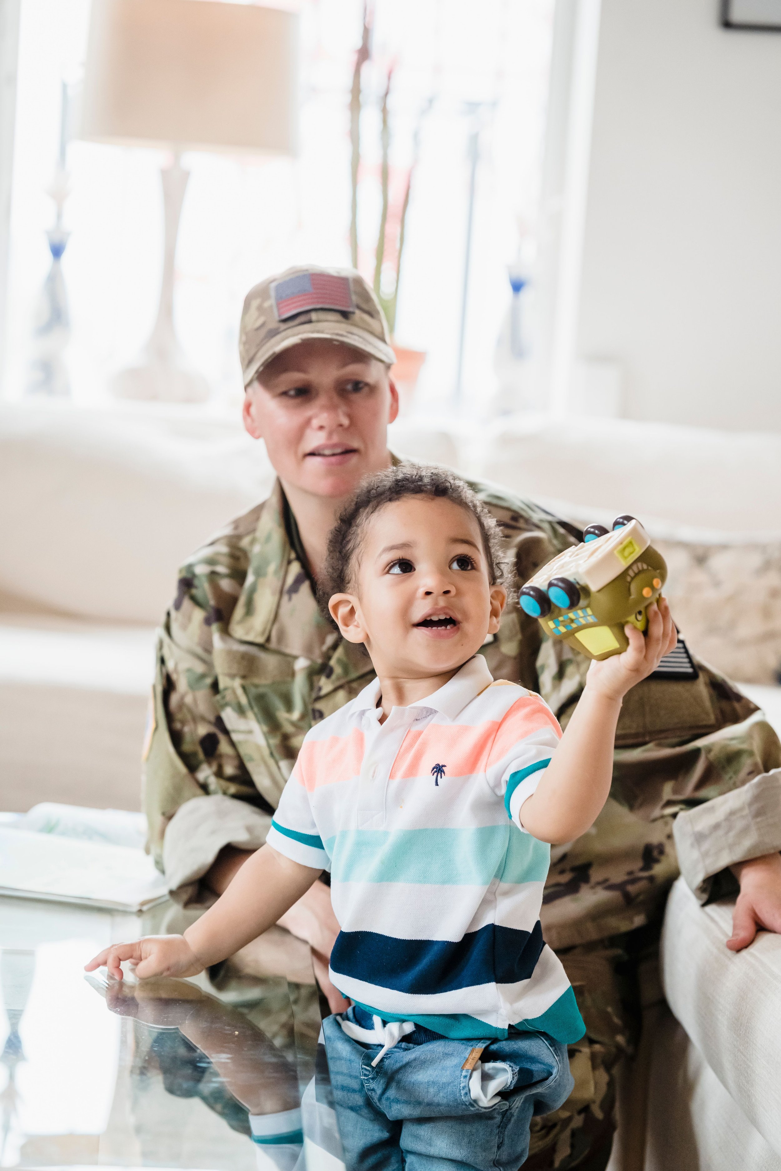 A soldier in camouflage uniform and cap with an American flag patch is sitting on the floor with a young boy. The boy is holding a toy and has a big smile. They are indoors, near a table with a reflective surface.