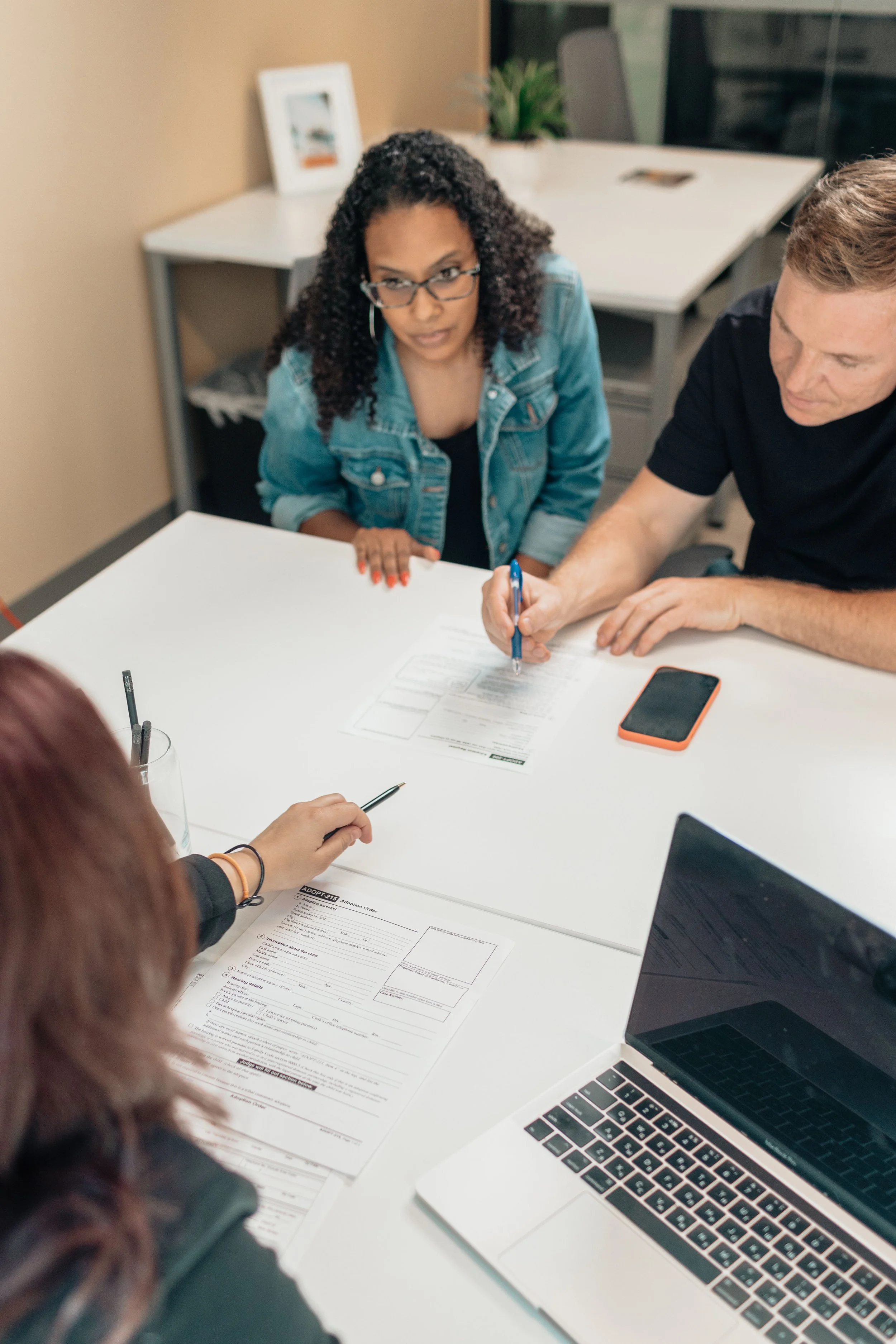 Two people are working at a white table with documents, laptops, and a smartphone. One person, wearing glasses and a denim jacket, is looking at the camera. The other person is writing on a document.