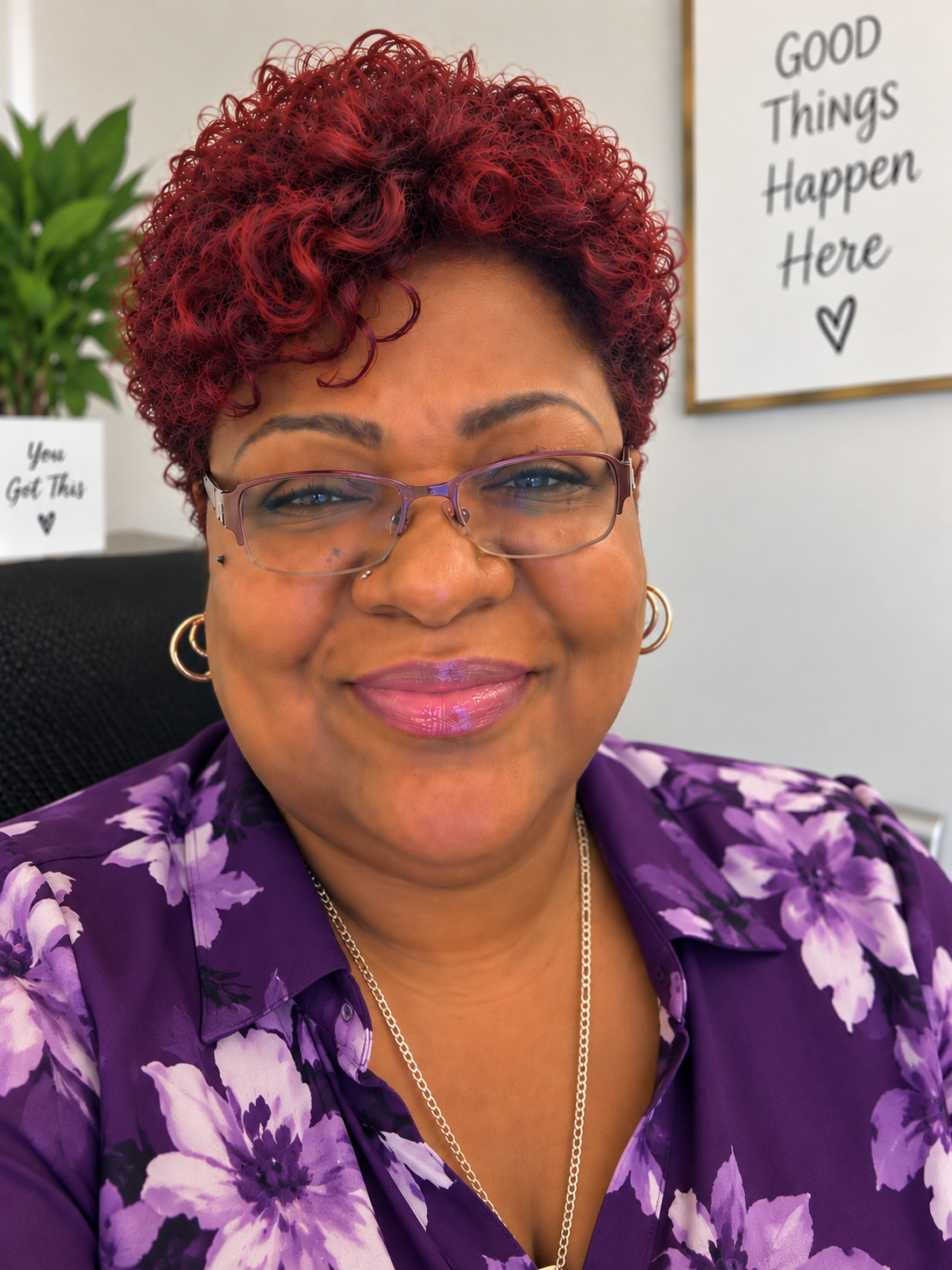 Close-up of a smiling woman with short, curly, red hair wearing glasses, gold hoop earrings, and a purple floral blouse. In the background, there are motivational signs and a green plant.