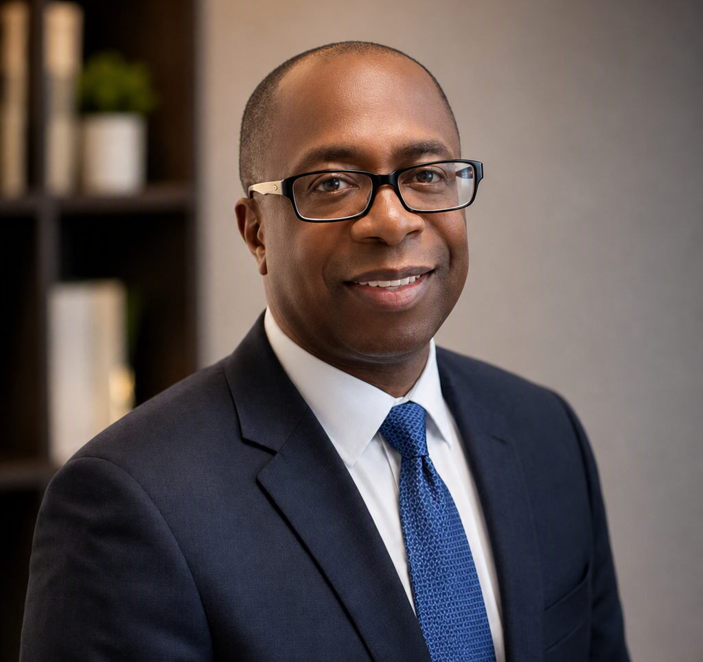 A professional man with glasses wearing a suit, white shirt, and blue tie, smiling in an office setting.