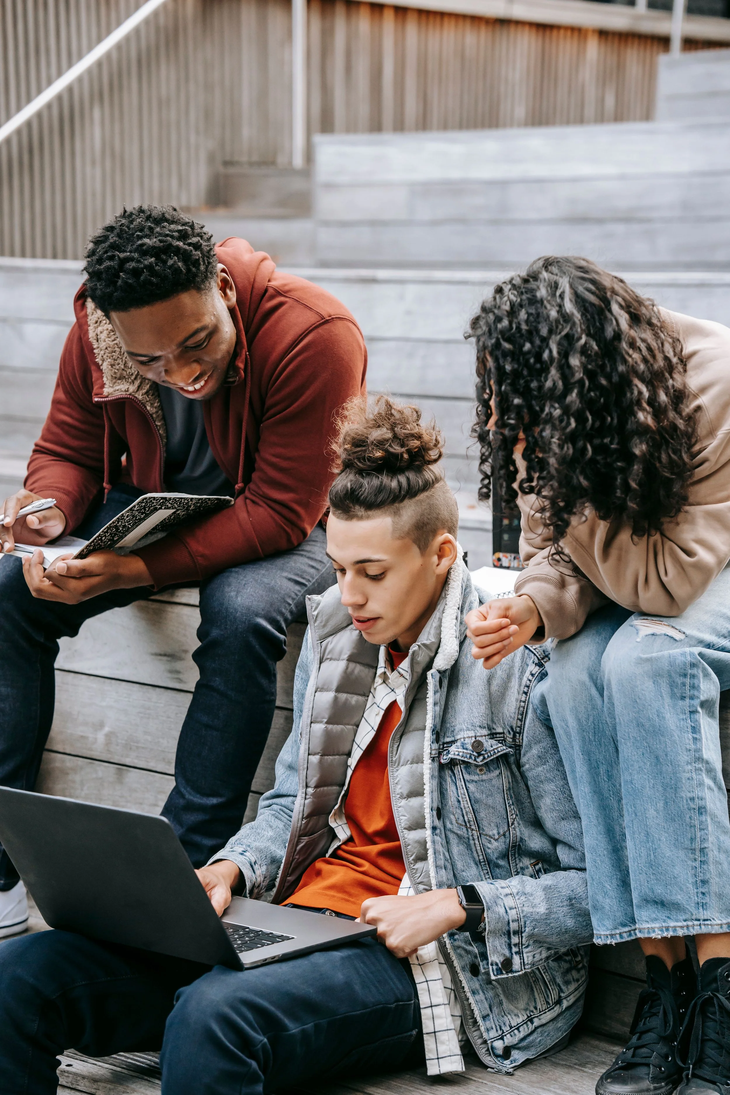 Group of four young people sitting on wooden stairs outside, engaged with laptops and phones, appearing to work or study together.