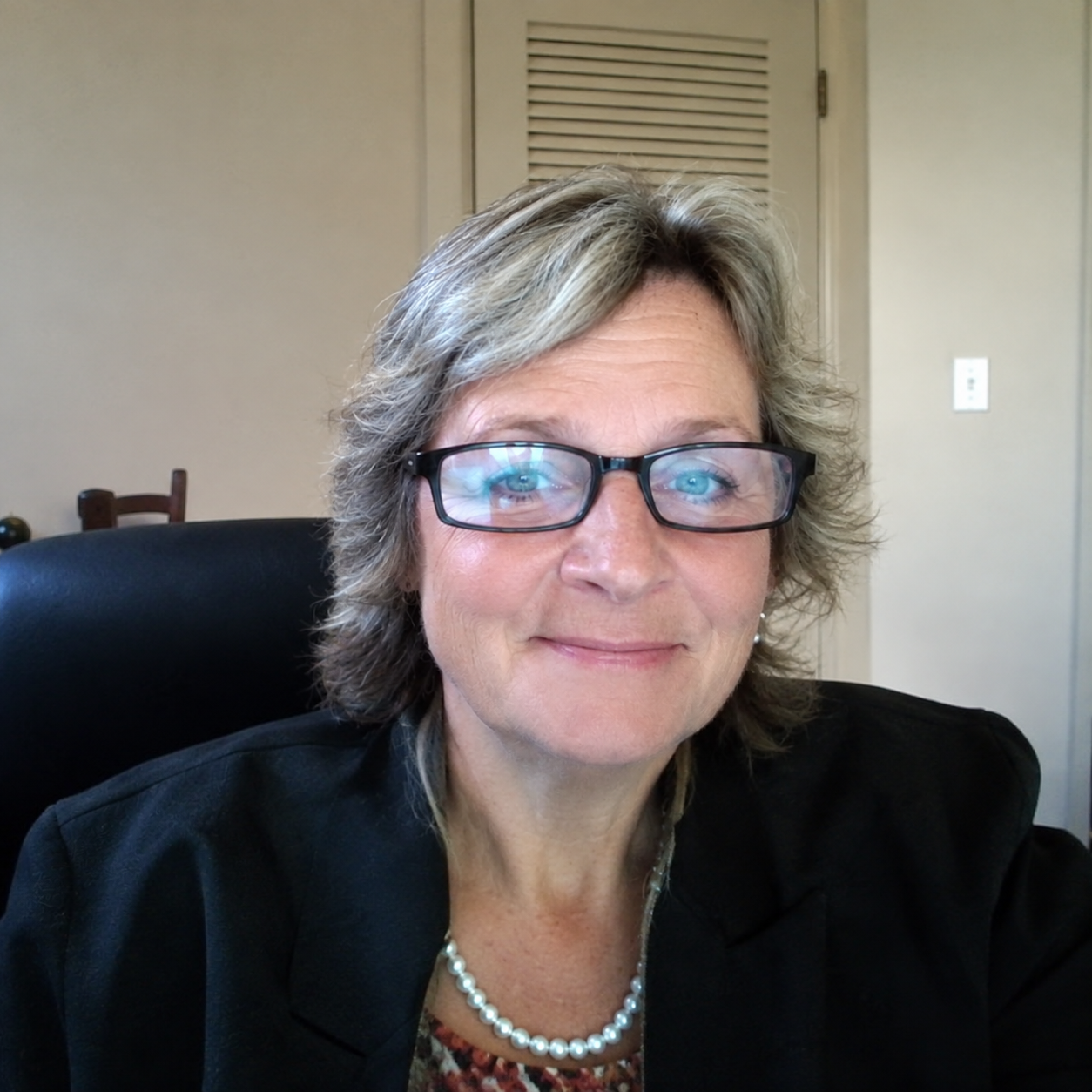 A middle-aged woman with short, wavy, gray hair and glasses, sitting in a black office chair, smiling at the camera with a beige wall and closed door in the background.