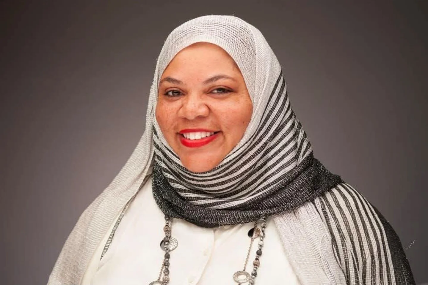 Portrait of a woman with a striped black and white headscarf, smiling with red lipstick, against a gray background.