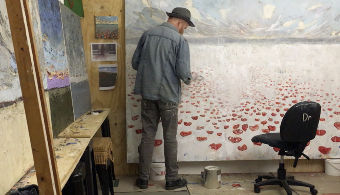 Artist painting a landscape of red poppies on a large canvas in an art studio, with a black chair labeled 'Dr', paint supplies, and art pieces on the wall.