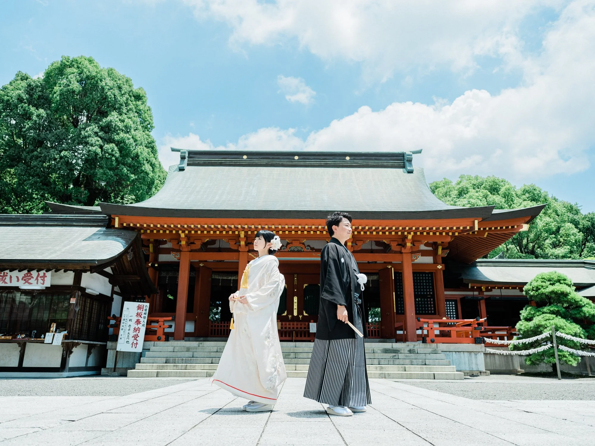 神社の前で着物と袴を着た男性と女性が並んで立っている。背景には青空と緑の木々が見える。