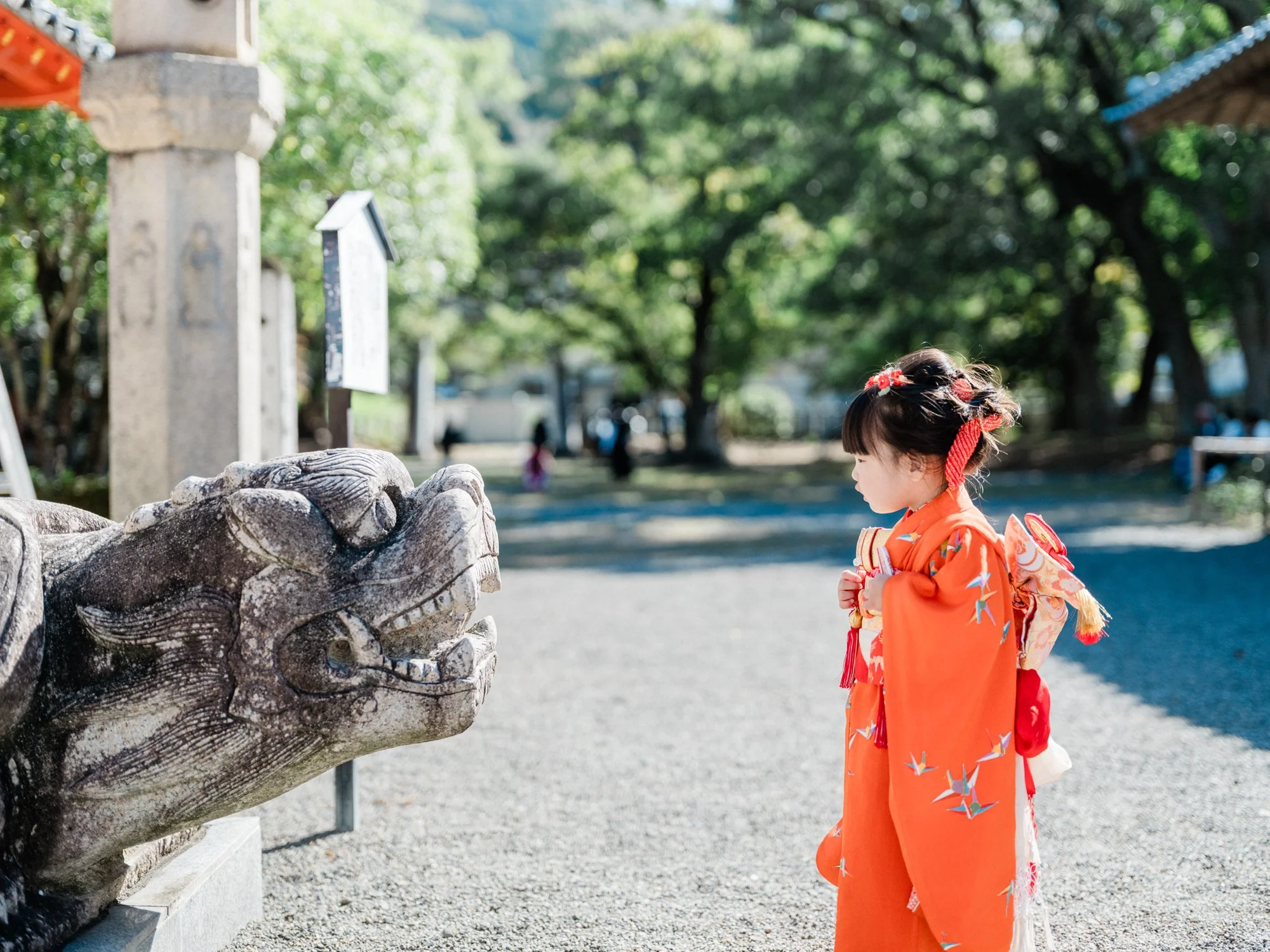 七五三の写真。伝統的な着物を着た少女が神社の石の狛犬像にお辞儀している様子で、背景には緑の木々と青い屋根の建物がある風景です。