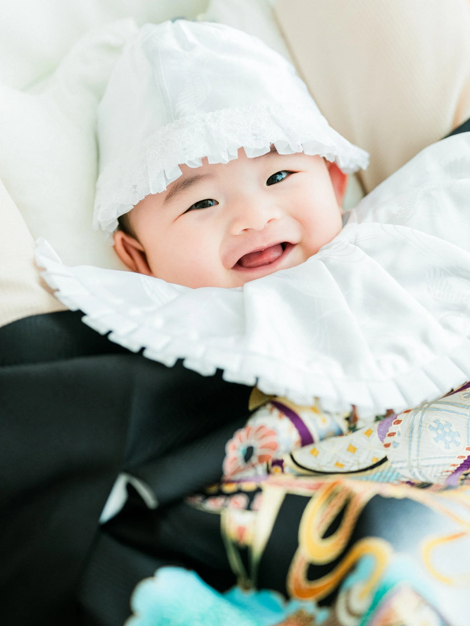 笑顔の赤ちゃんが白い帽子をかぶり、産着を着ている。お宮参りの写真。