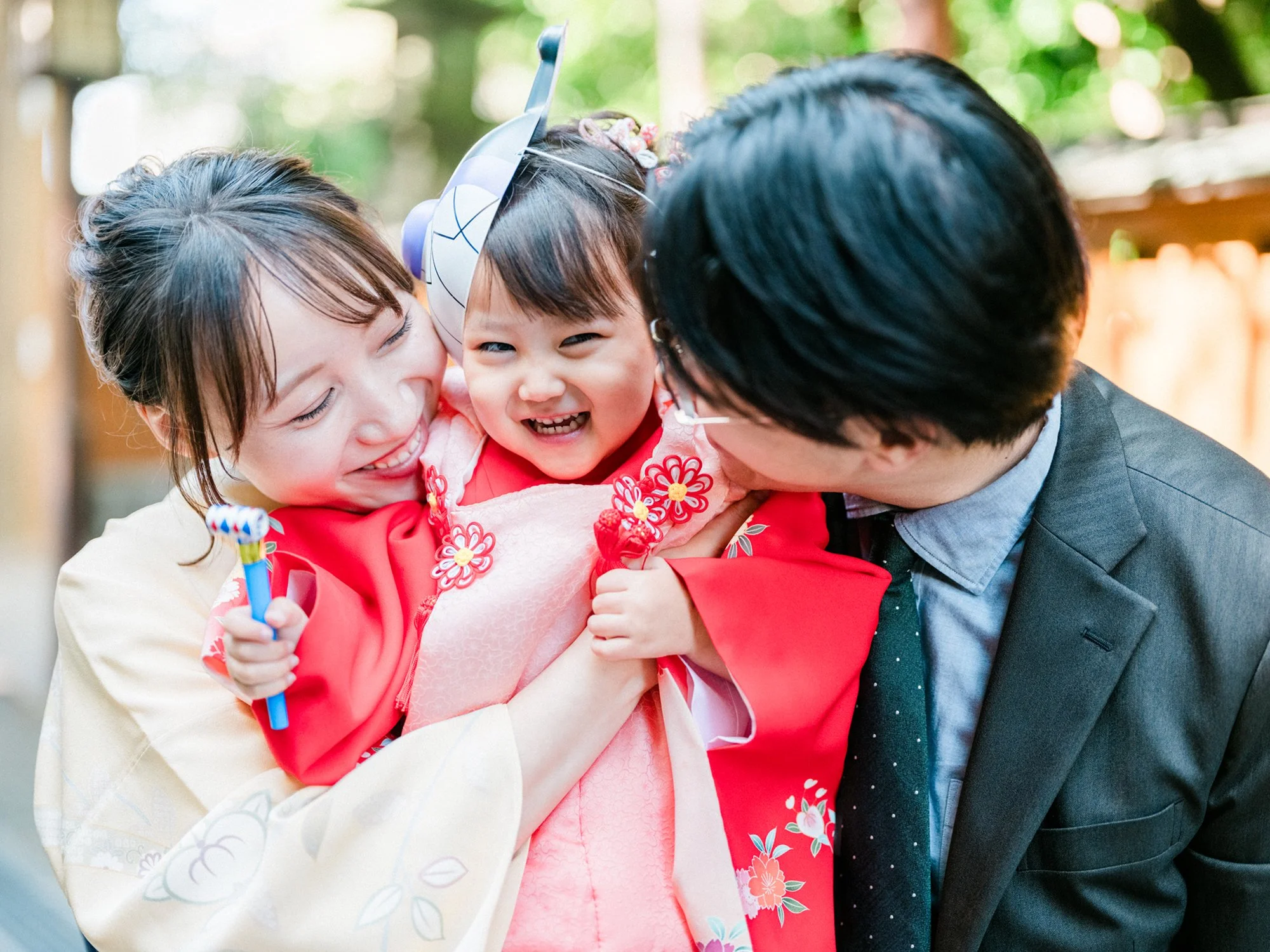 三人の大人と一人の子供が抱き合いながら笑顔で楽しんでいる写真。子供は赤い着物を着ており、頭に飾りをつけている。背景は緑豊かで自然光が差し込んでいる。