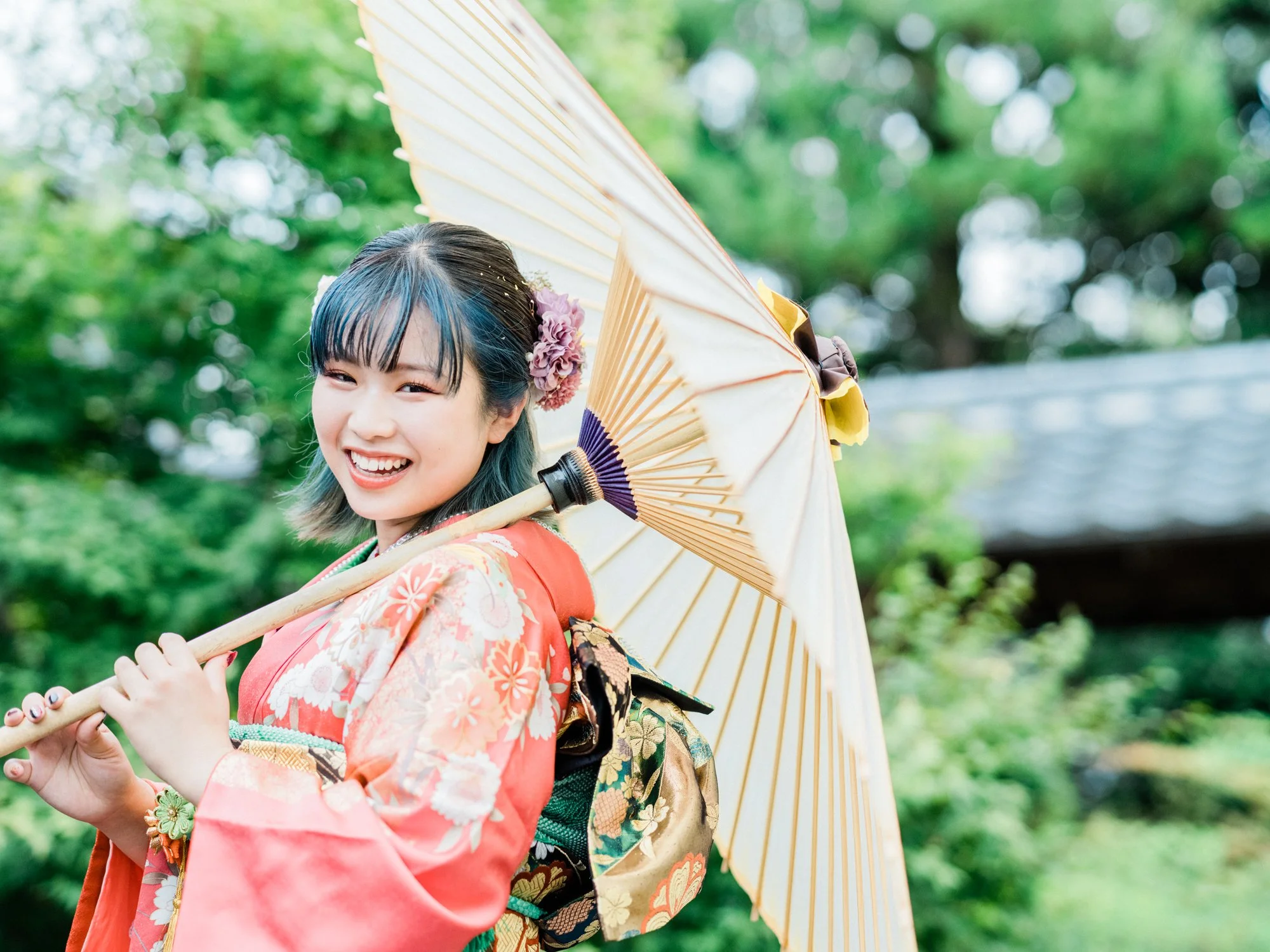 振袖を着た女性が和傘を持ち、笑顔で振り向いている。背景は緑の木々です。ナチュラルな成人式の前撮り。