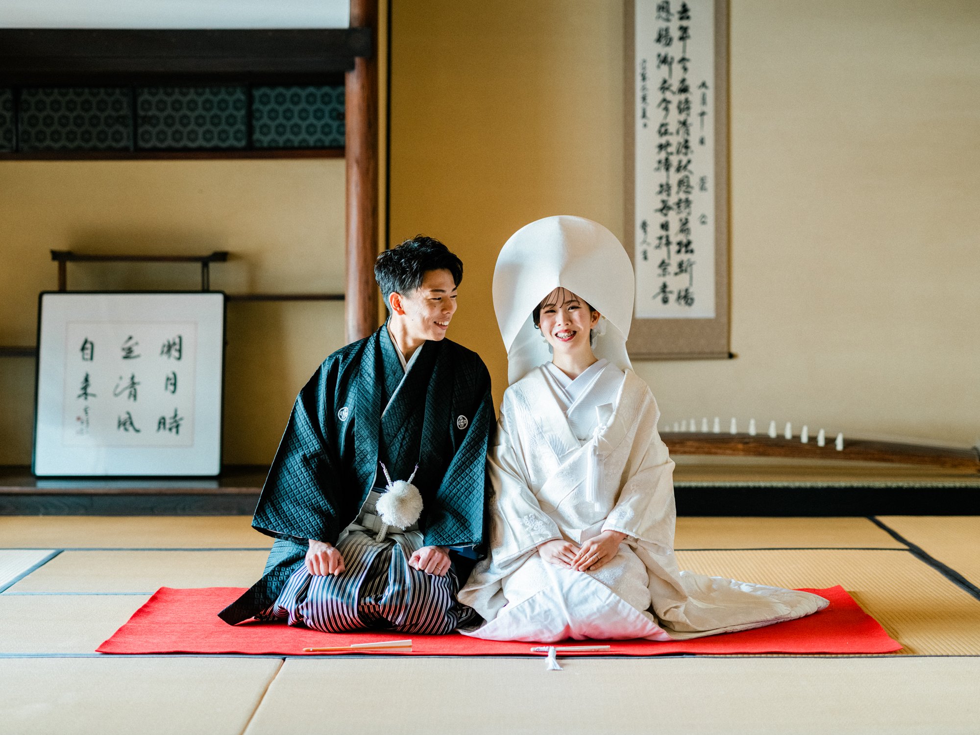 伝統的な和装をした新郎と白無垢を着た新婦が座って笑顔で並んでいる。背景には和室と掛軸が見える。古民家での結婚式前撮り。