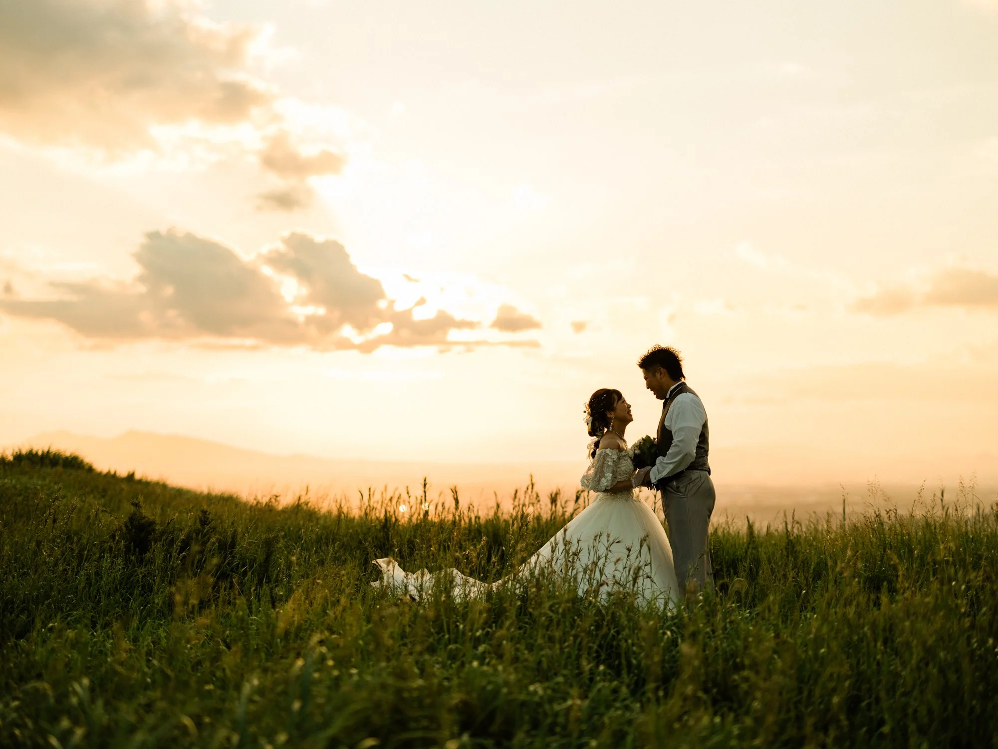 夕焼けの空の下で結婚式を挙げるカップル。草原に立ち、互いに見つめ合っている。