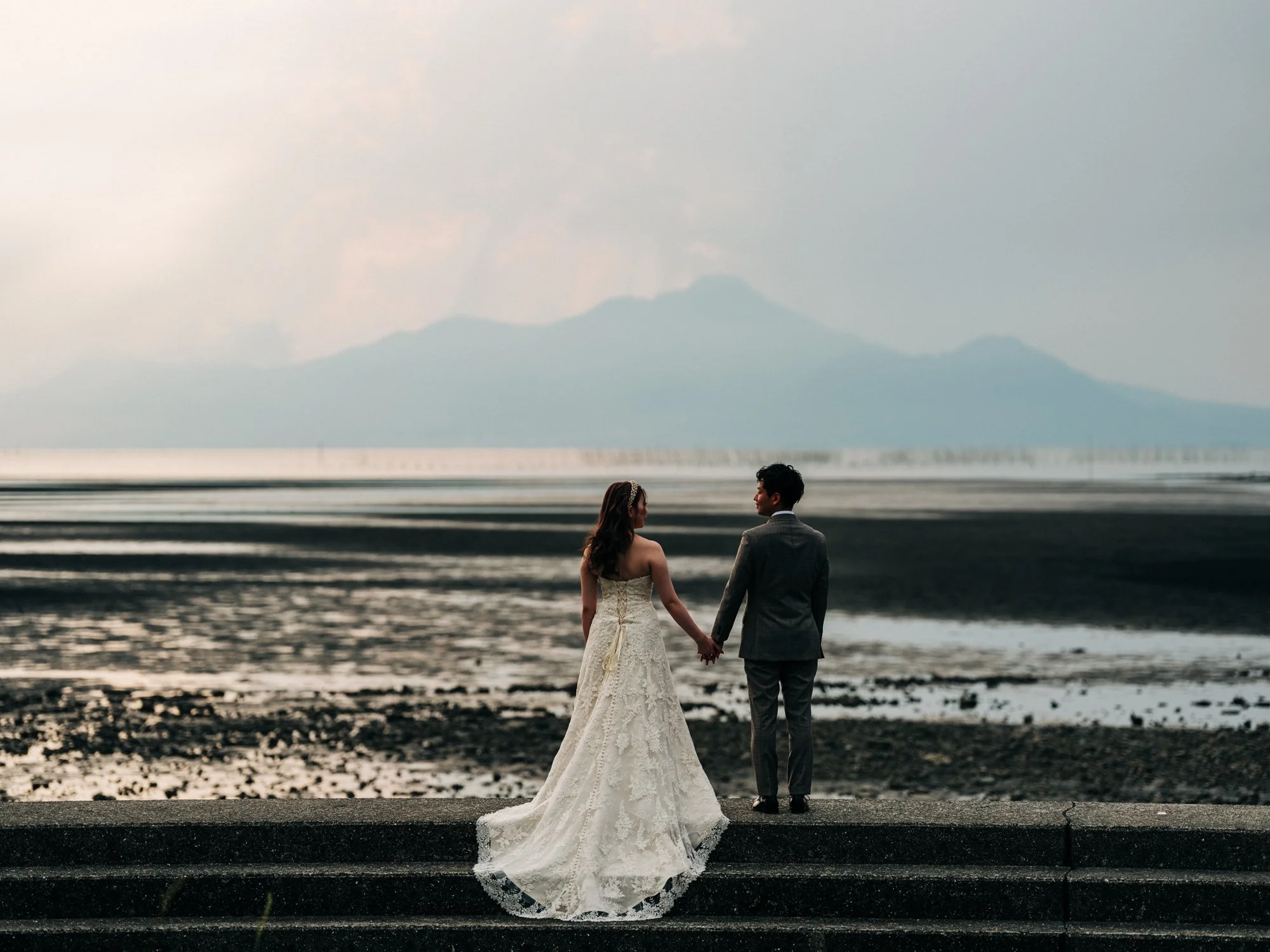 海辺で結婚式を挙げる新郎新婦が手をつないでいる。背景には山と空が広がっている。