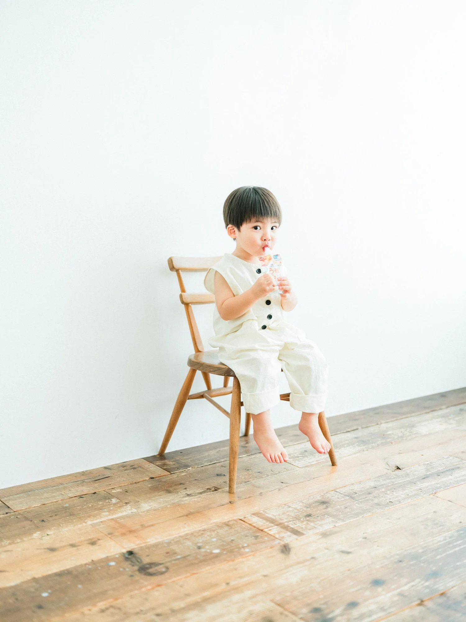 白い背景の前に座っている幼い男の子が、ゼリーを飲んでいる。木製の椅子に座っており、素足で木の床の上にいる