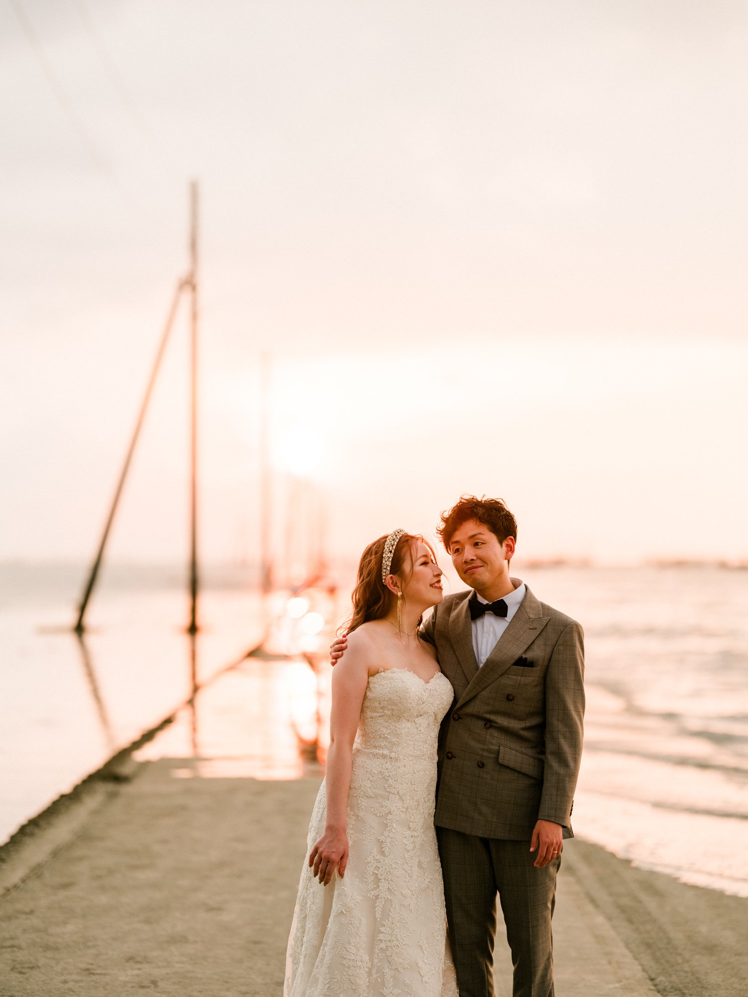 夕焼けの海辺でウェディングドレスの花嫁とスーツの花婿が抱き合っている。