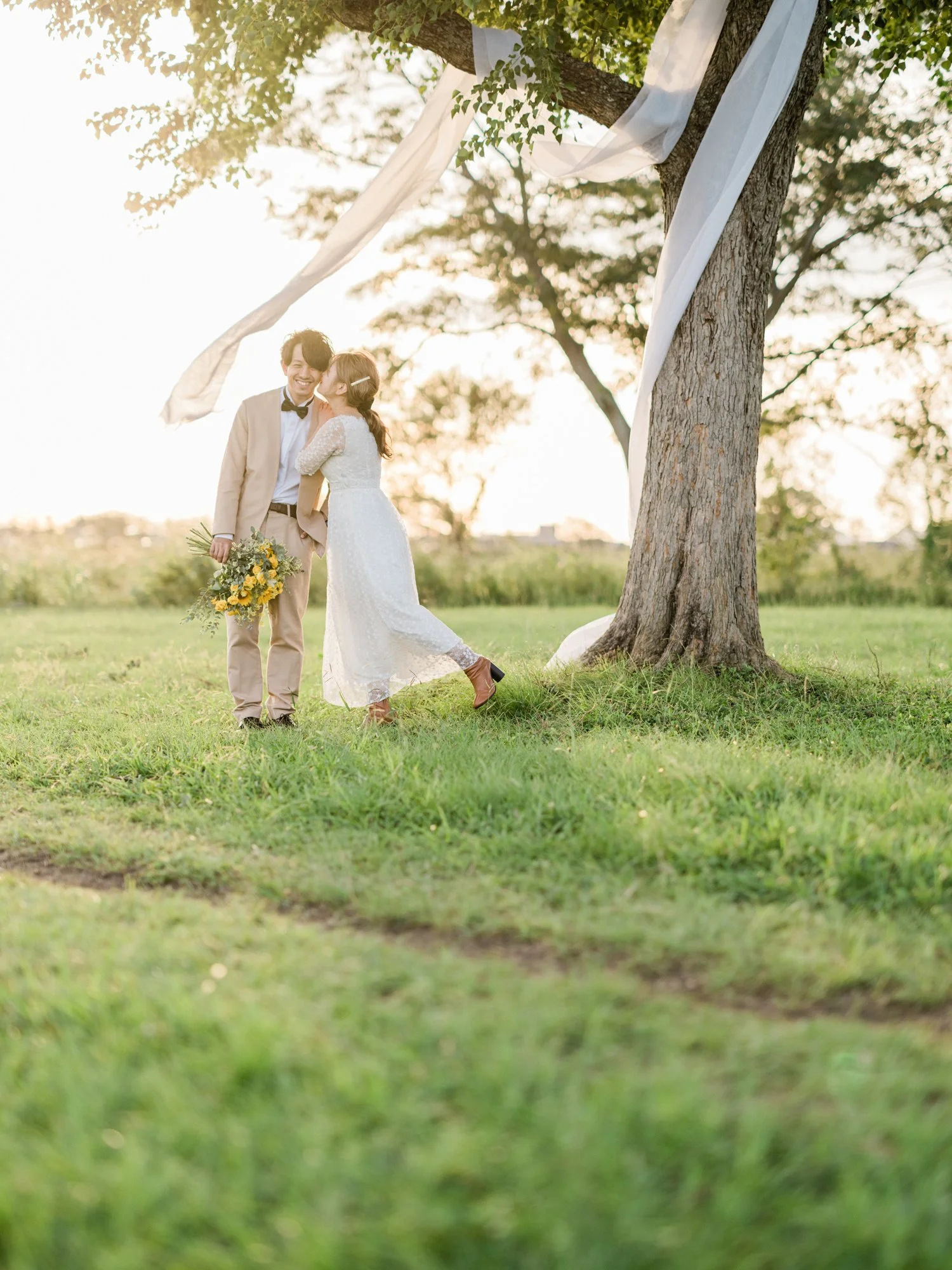 木の下で結婚式をしているカップル。女性は白いドレス、男性はベージュのスーツと蝶ネクタイを着ており、女性は花束を持っている。夕暮れ時らしき暖かい光の中で撮影されている。