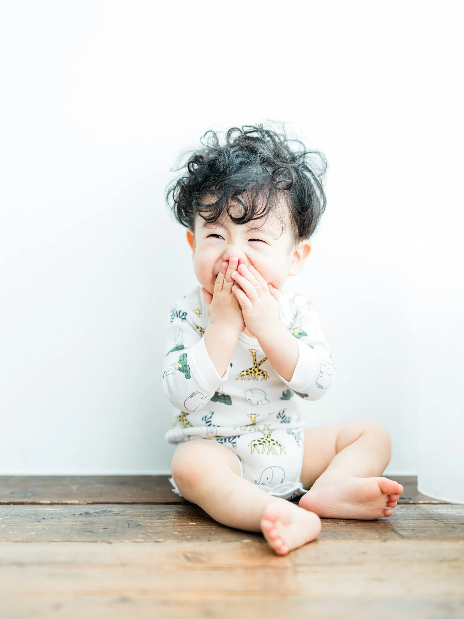 笑っている赤ちゃんが座っている写真。白い背景と木の床の上に座っており、動物柄の洋服を着ている。