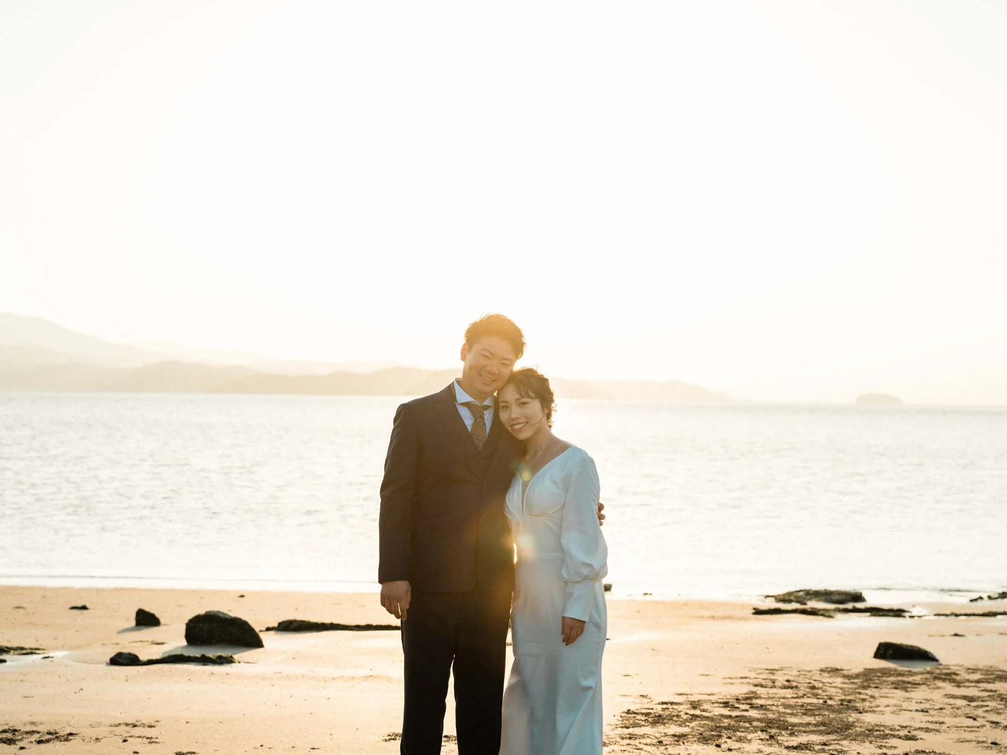 日没時の海辺で笑顔の夫婦が手をつないでいる結婚式前撮り写真。