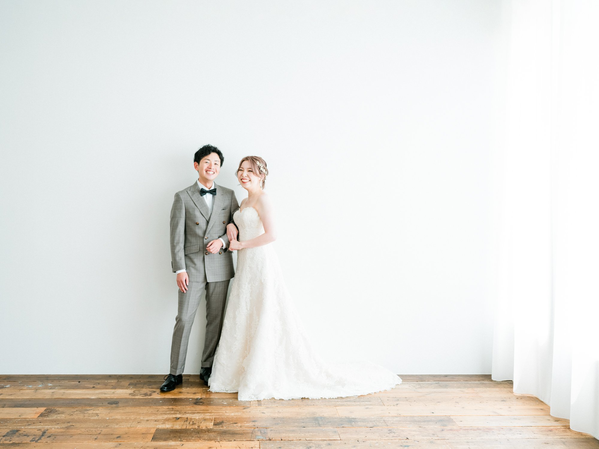 結婚式の新郎新婦のカップルが笑顔で並んでいる。新郎はグレーのスーツ、蝶ネクタイを着用し、新婦は白のドレスを着ている。白い壁の前でポーズを取っている。