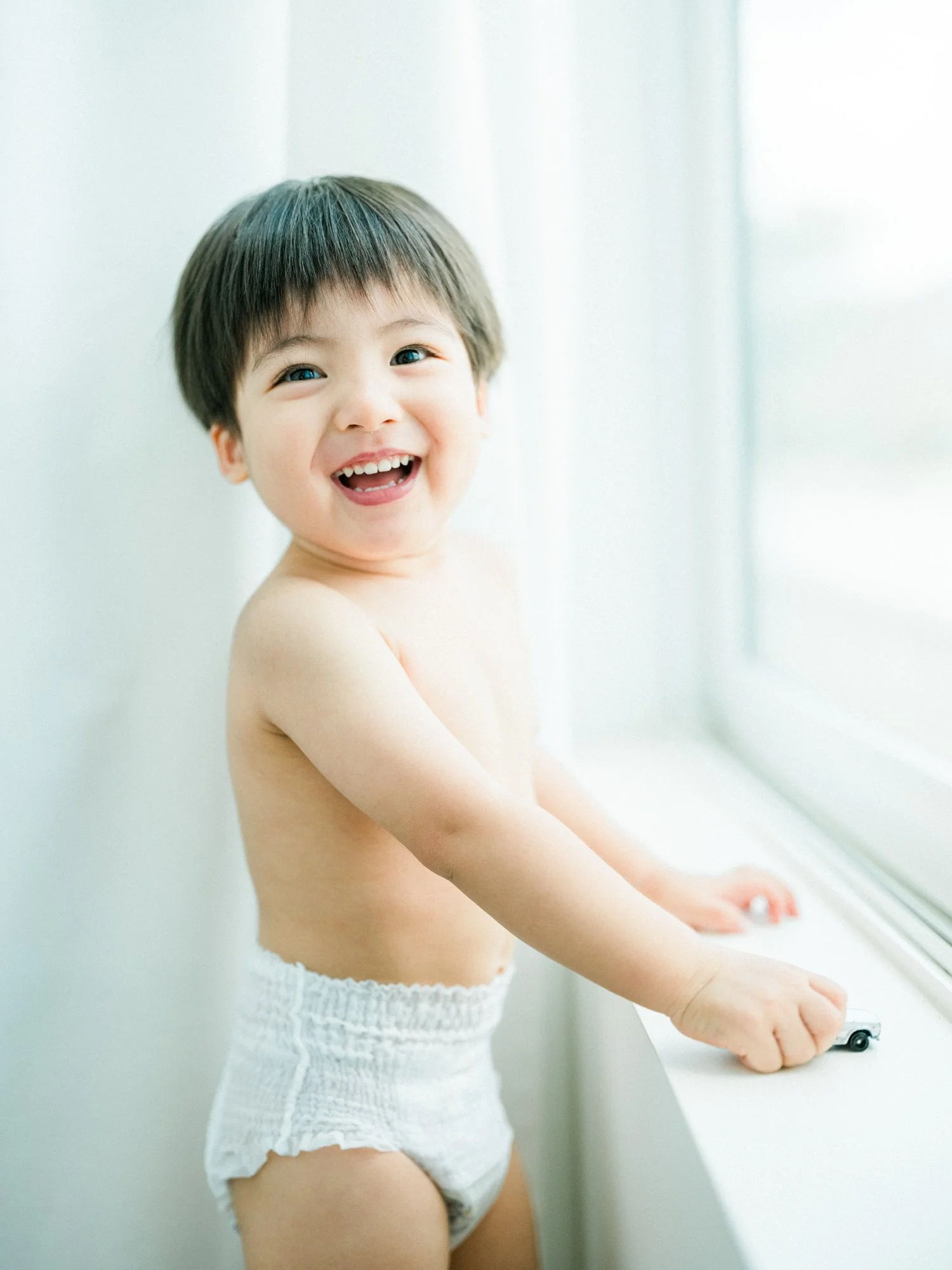 幸せそうに笑う幼い男の子が白いパンツを履き、窓辺で玩具の車を持っている