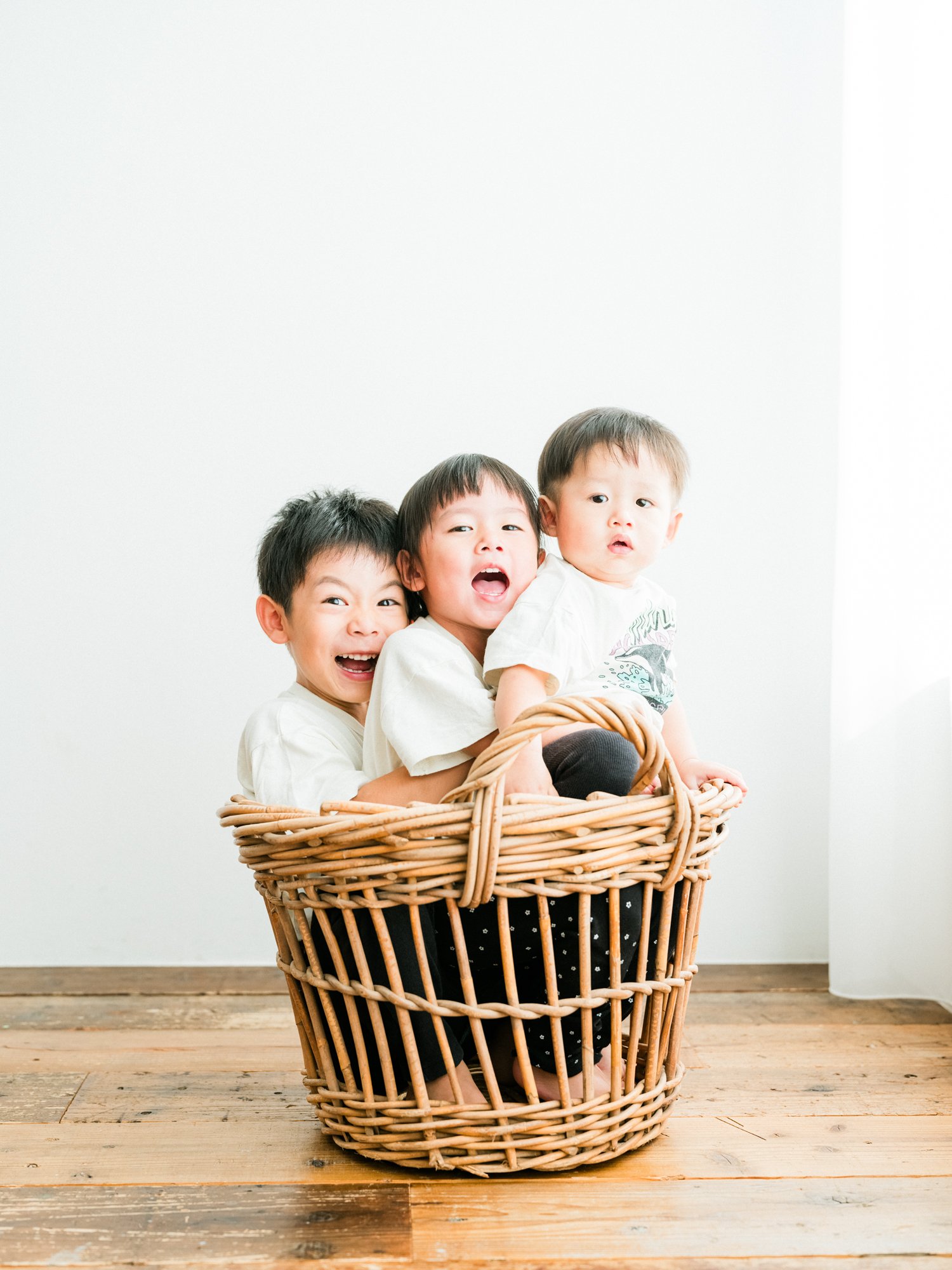 3人の子供が籠の中に入っている写真。子供たちは笑顔と驚きの表情をしている。背景は白い壁と木の床。