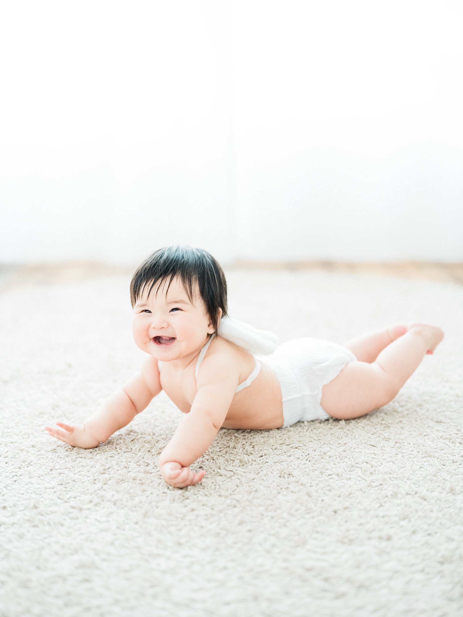 笑顔の赤ちゃんが寝転んでいる画像。赤ちゃんは白い衣服を着ていて、背景はラグの上にいます。