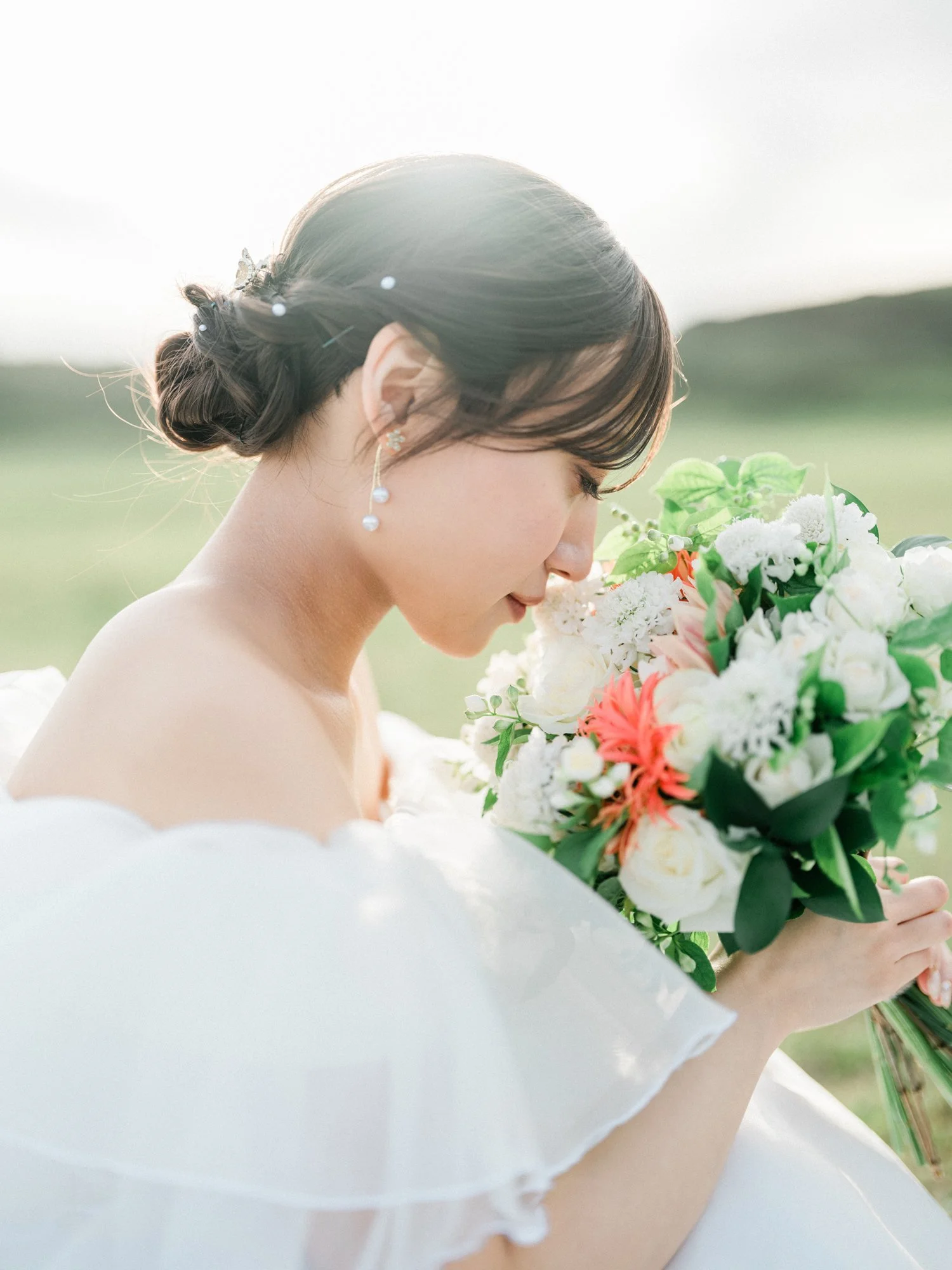 結婚式の花束を持つ女性の横顔。髪はアップにまとめており、イヤリングをつけている。背景は自然の風景で、柔らかな光に包まれている。
