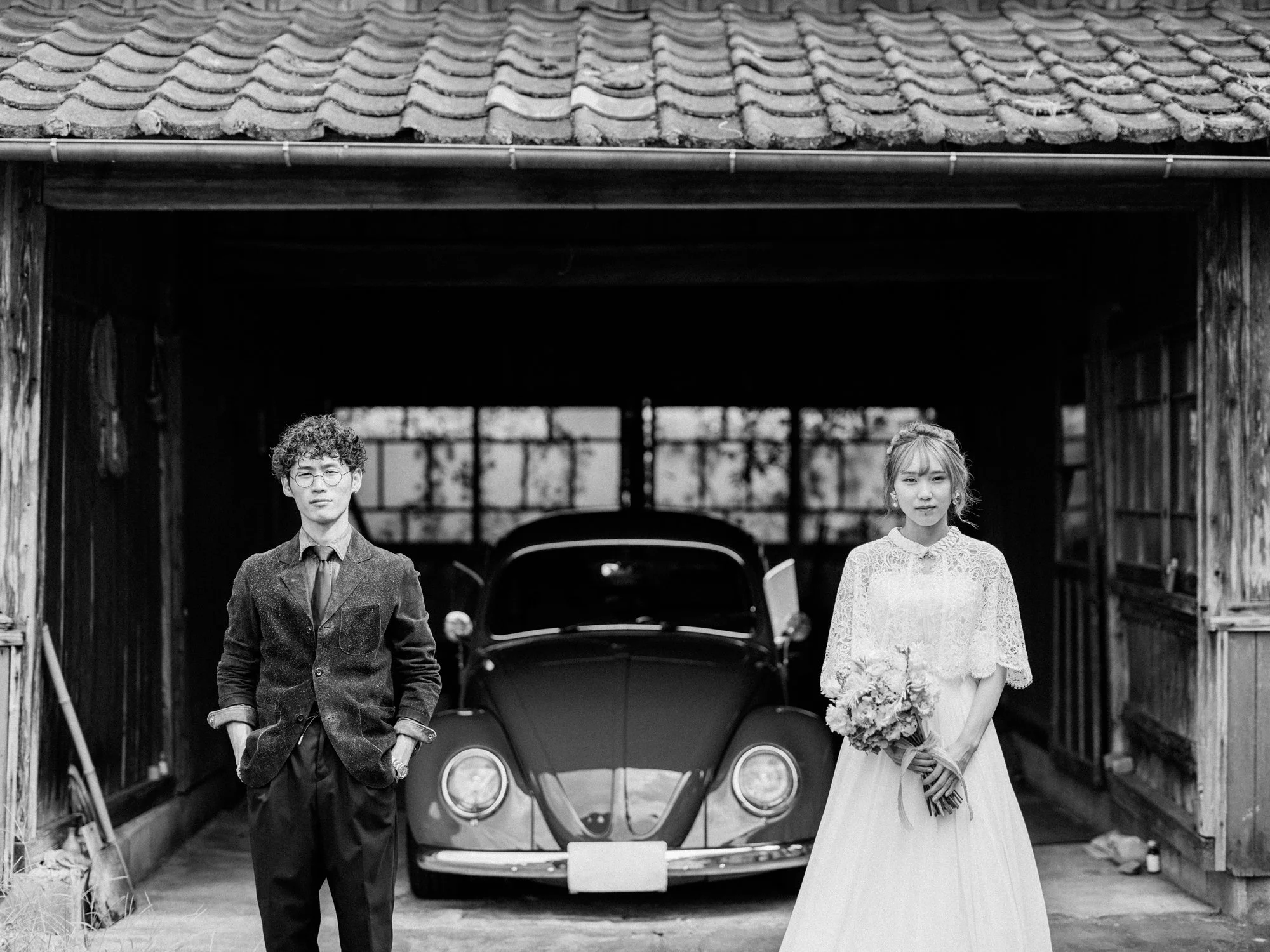 木造の建物の前に、男性と女性が結婚式の衣装を着て立っている。背景には車があり、屋根は瓦屋根で、木製の壁とゲートがある。