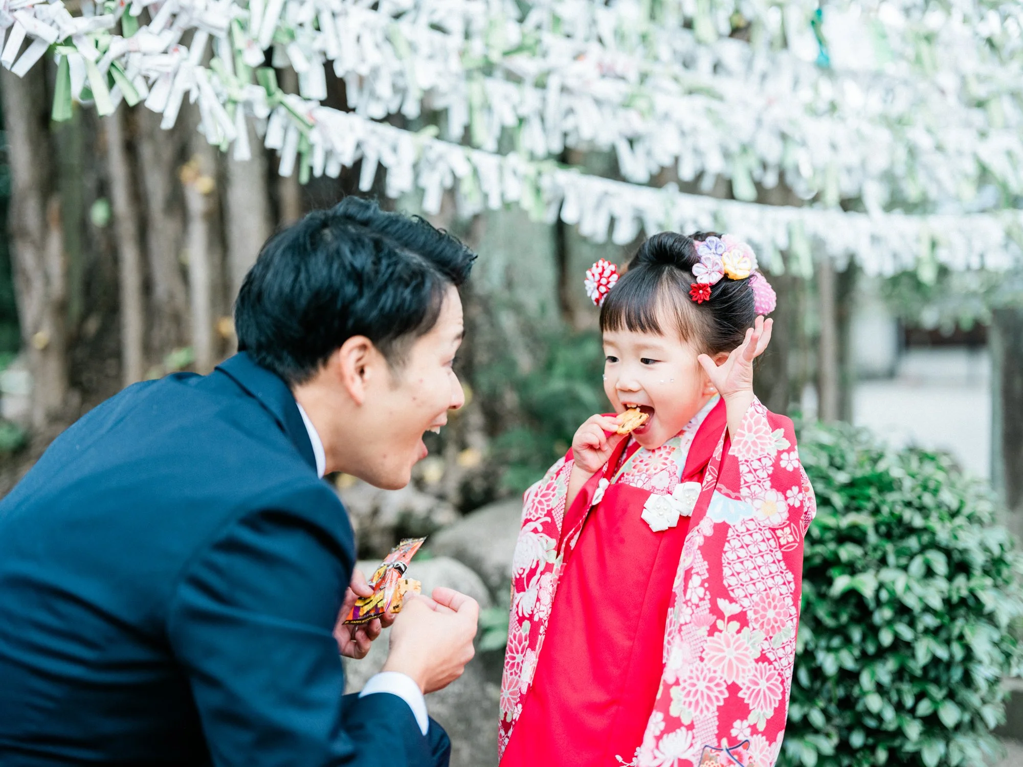 若い女の子が赤い着物を着て髪には花の髪飾りをつけ、笑顔で手にお菓子を持ちながら、大人と会話をしている。