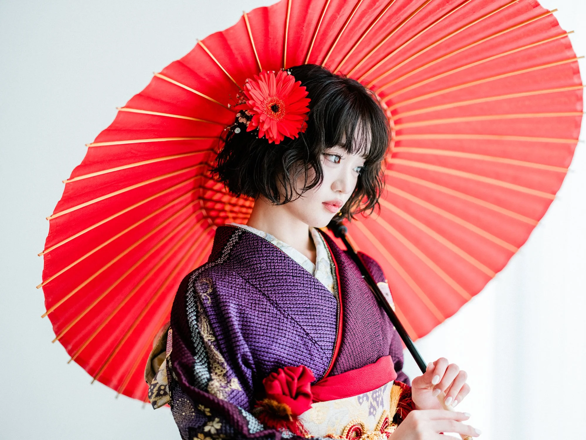 振袖を着用し、赤い髪飾りをつけた女性が赤い紙の傘を持ち、横を向いている。