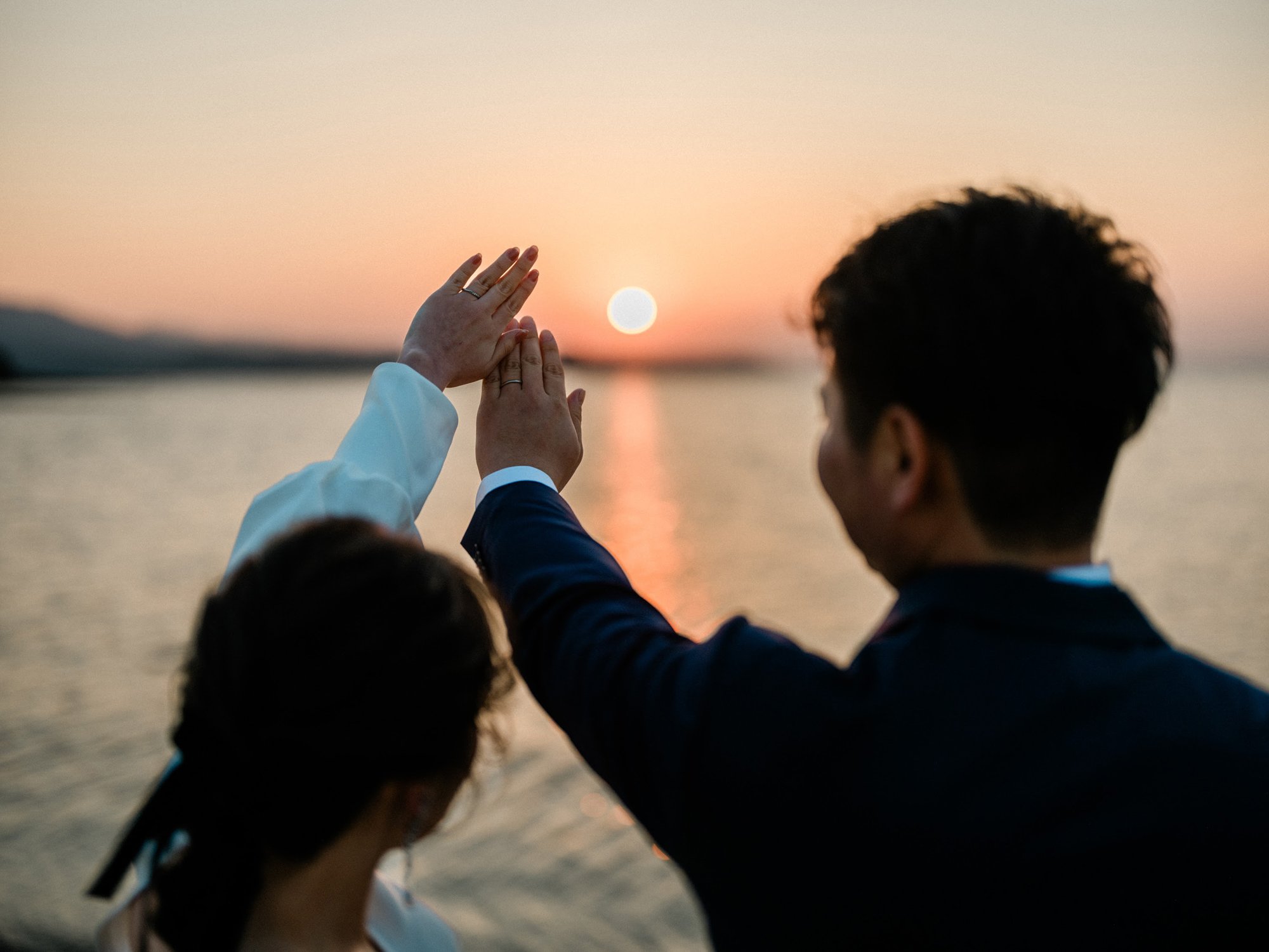 夕日に向かって新郎新婦が指輪を見せている光景。