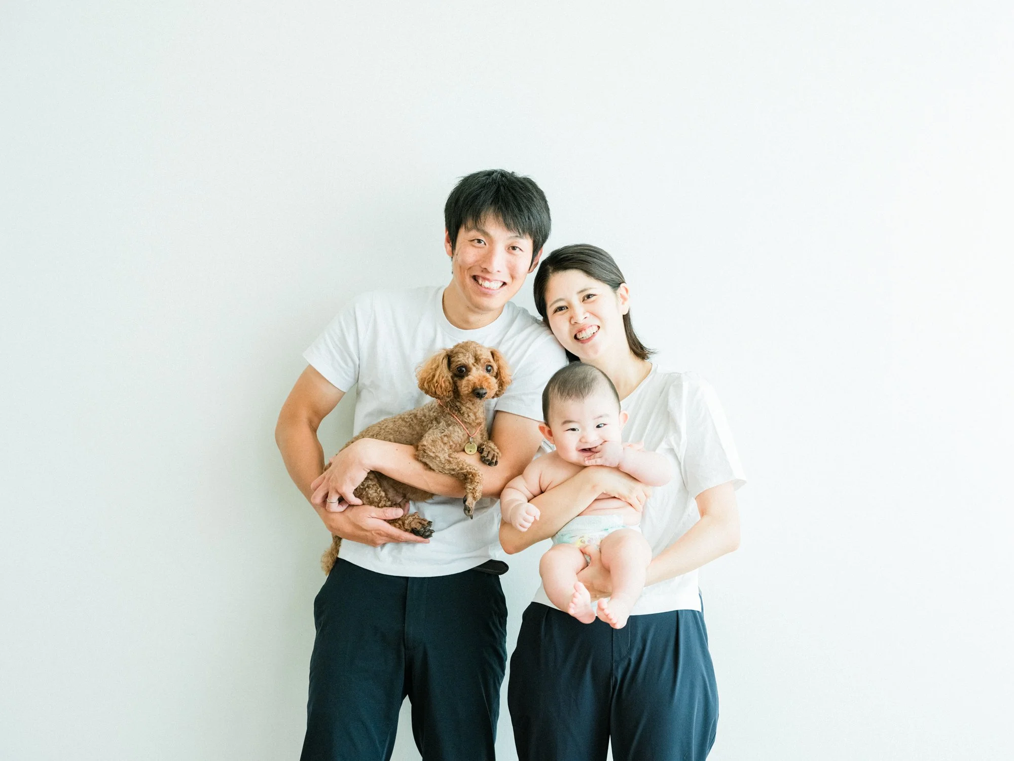 白い背景の前で笑顔の若い夫婦と子ども、犬を抱きしめている。