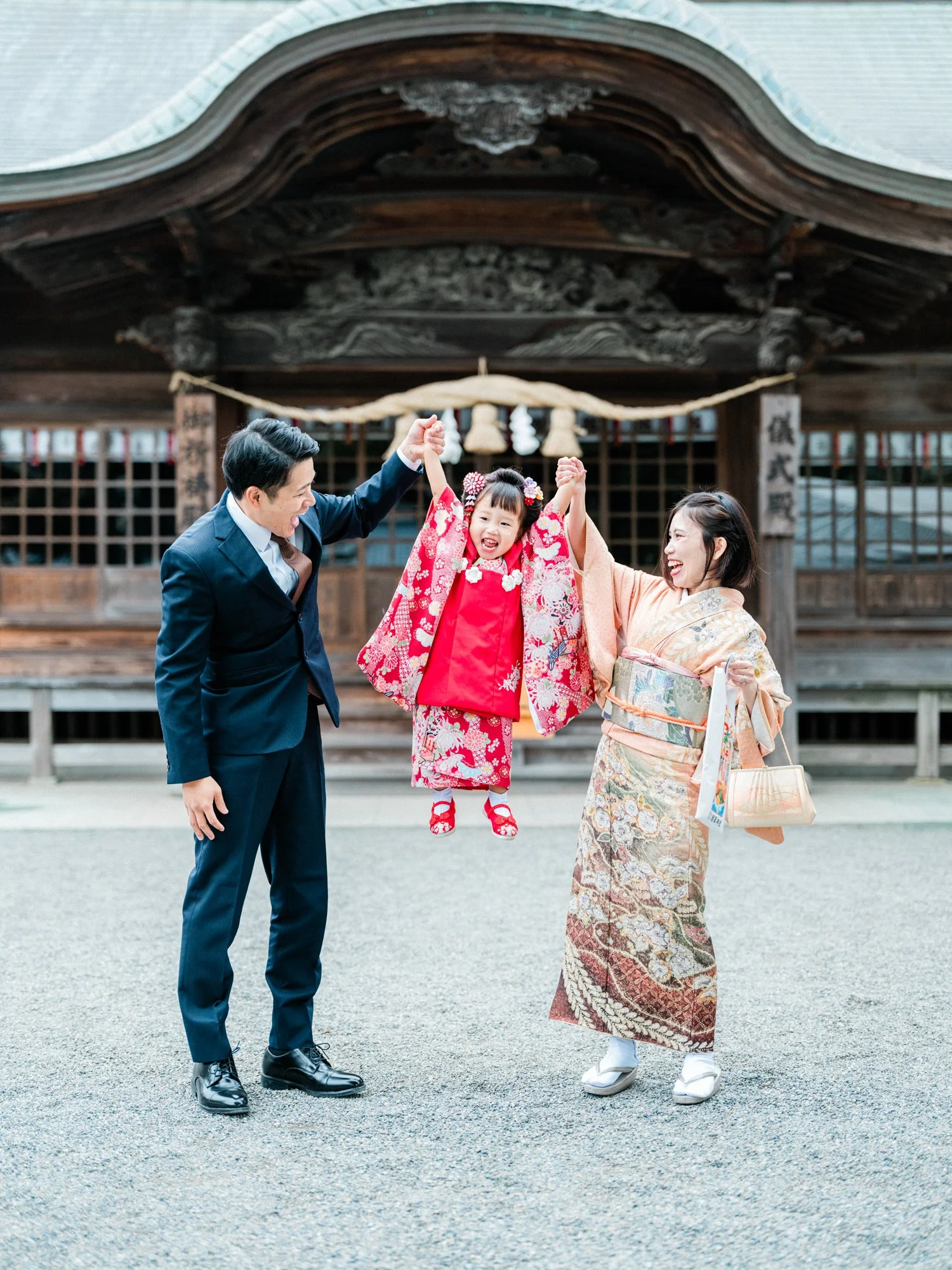 家族が神社の前でお祝いをしている。父親と母親が揺れる子どもを支え、笑顔で祝福している。子どもは伝統的な七五三の着物を着ている。
