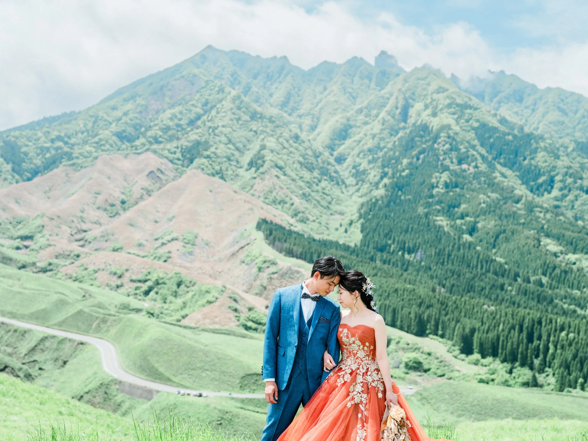 緑豊かな山々の背景に、ウェディングドレスを着た花嫁さんとタキシードを着た花婿さんが寄り添っている写真です。