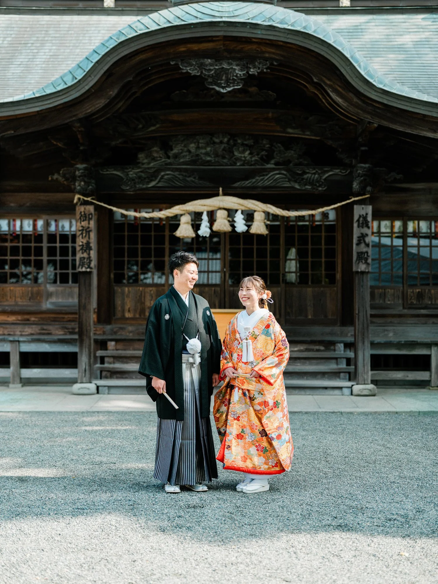 伝統的な着物を着た新郎新婦が健軍神社の前で笑顔で並んでいる。