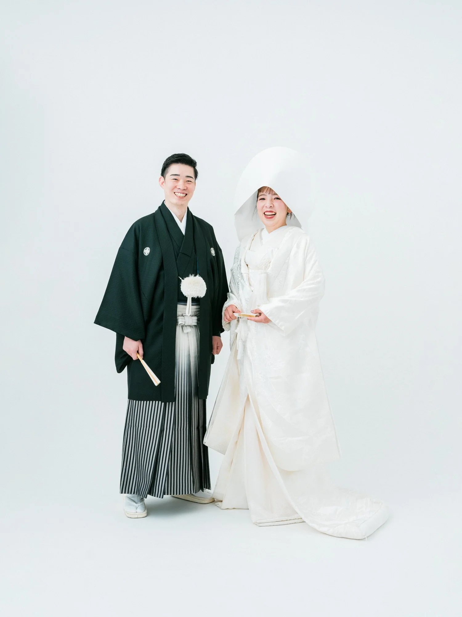 紋付袴を着た男性と白無垢、綿帽子の女性が幸せそうに笑顔で立っている写真。背景は白色でシンプルです。