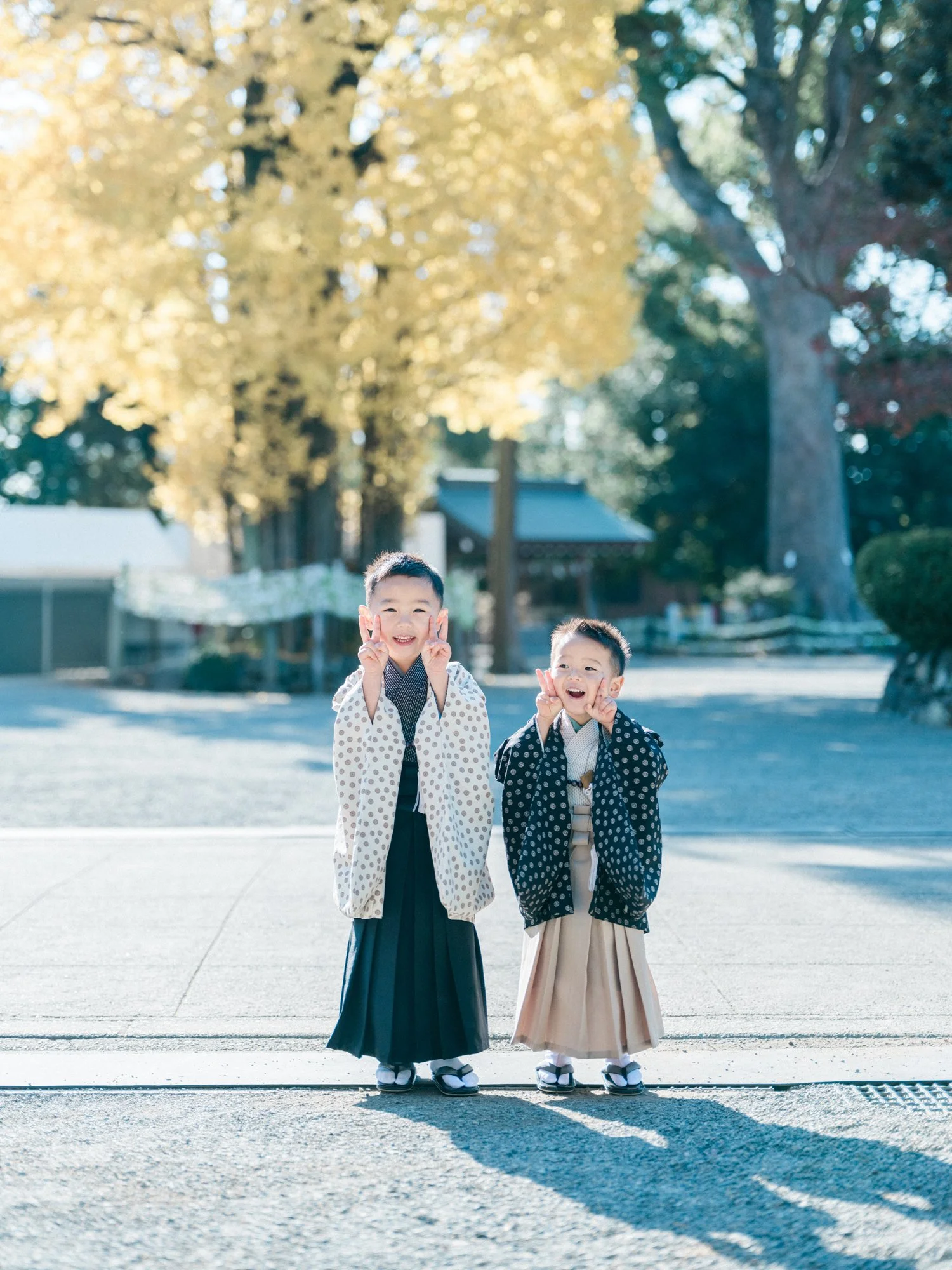 伝統的な七五三の着物を着た二人の子供が神社で笑顔でポーズを取っている。背景に大きな木と建物が見える青空の下の風景。