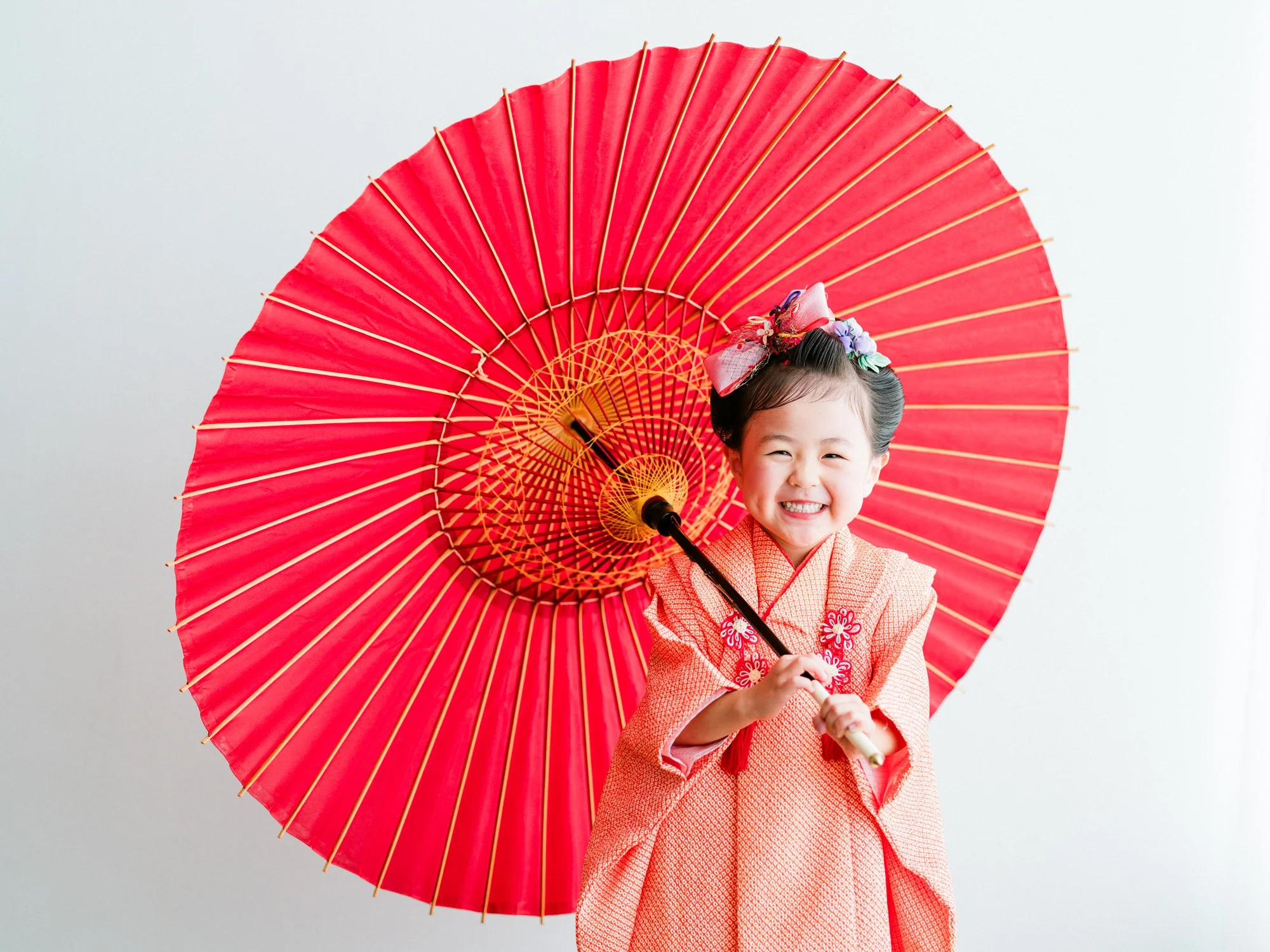 笑顔の子供が赤い着物を着て、赤い紙製の和傘を持っている。