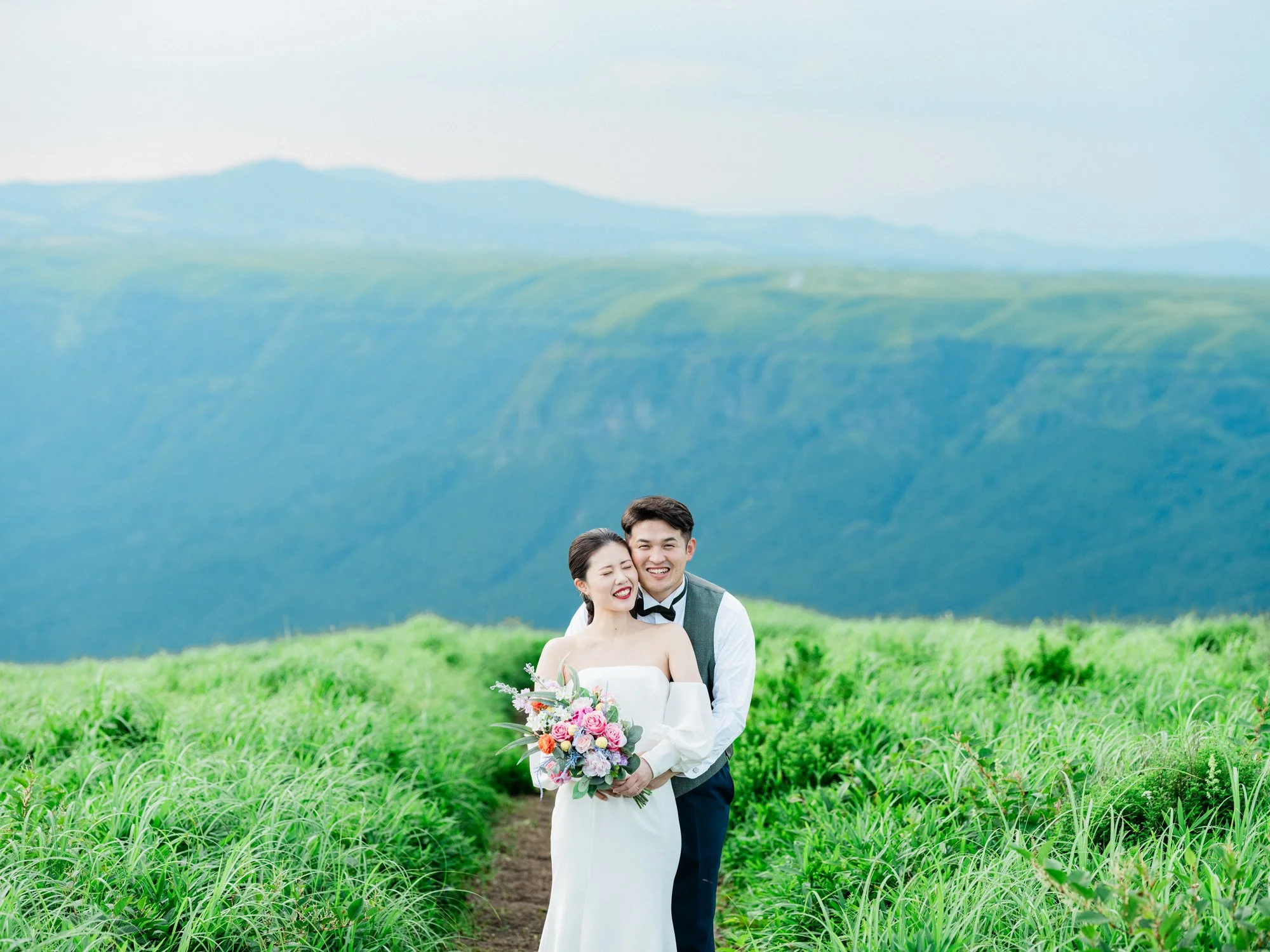 山の緑豊かな風景の中で、結婚式を挙げているカップル。花嫁は白いドレスと花束を持ち、笑顔でカメラを見ている。花婿は彼女の後ろに立ち、笑顔を見せている。