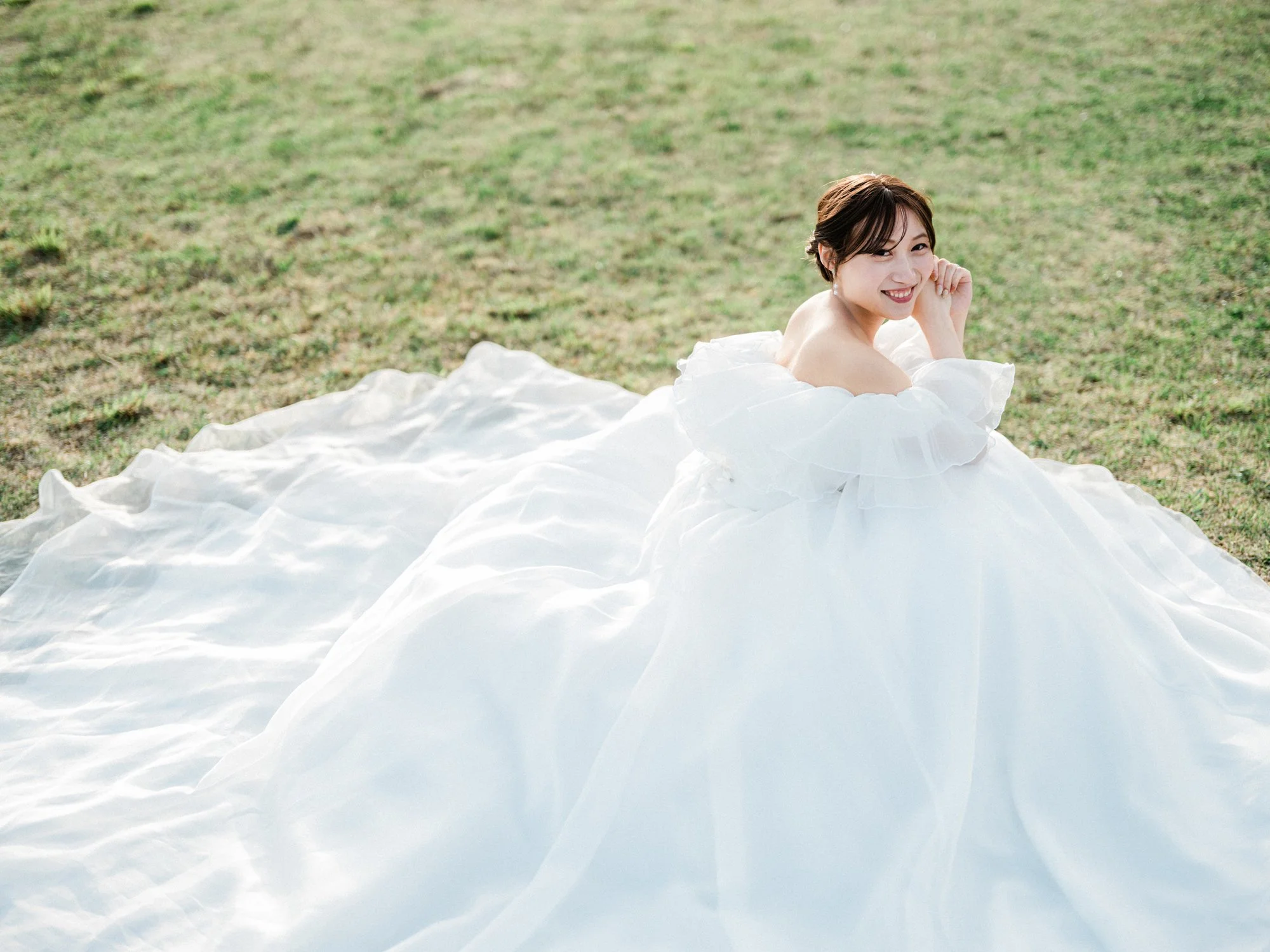 笑顔の女性が白いドレスを着て草原の上に座っている写真。