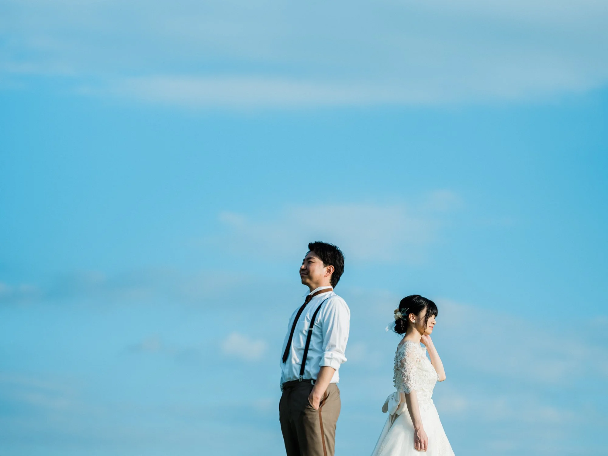 青空の下で向かい合って立つ結婚式のカップル。男性は白いシャツとネクタイ、女性は白いウェディングドレスと花の髪飾り。