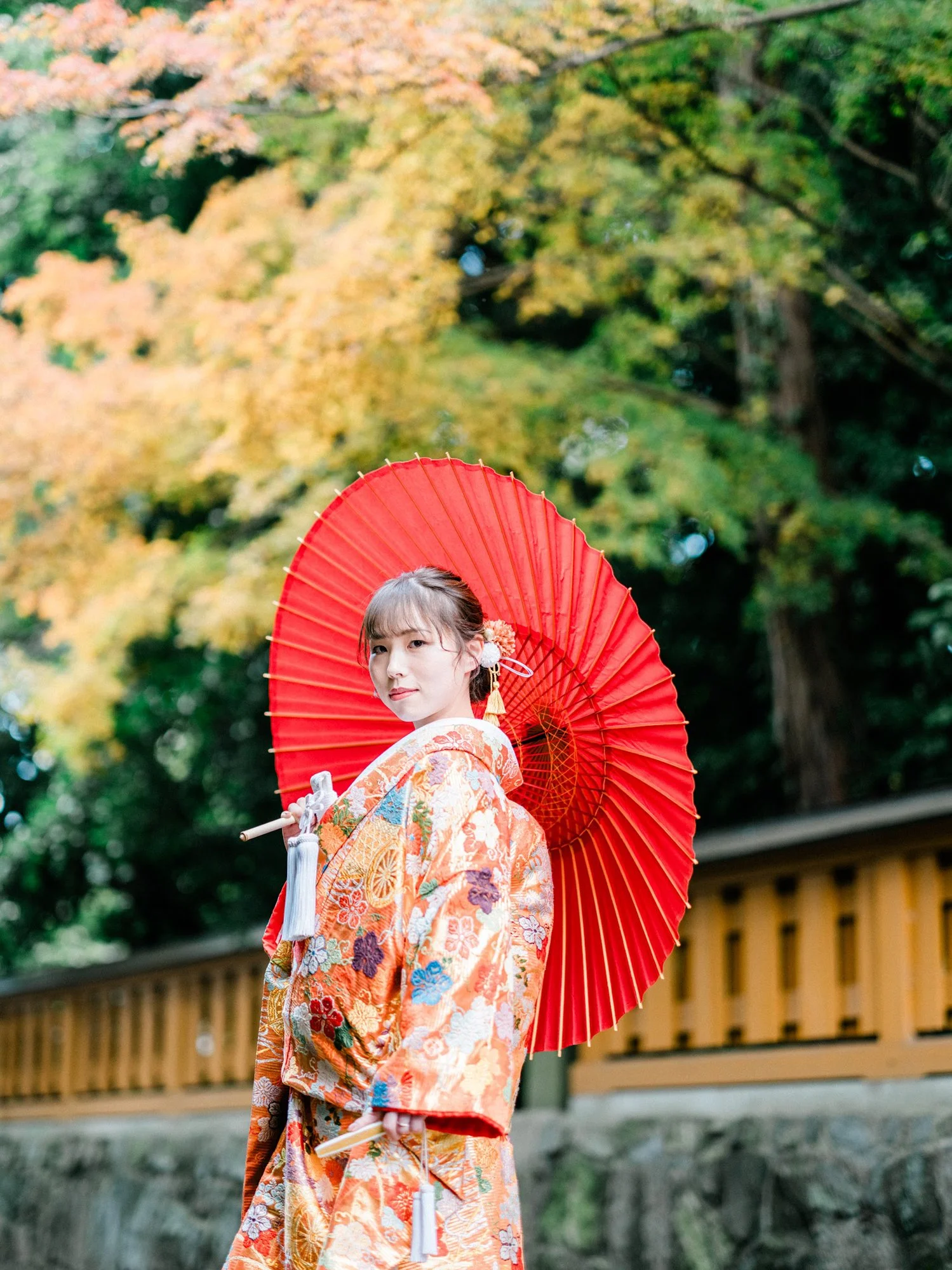 伝統的な着物を着て紅色の傘を持った女性が、秋の紅葉が背景の庭園に立っている