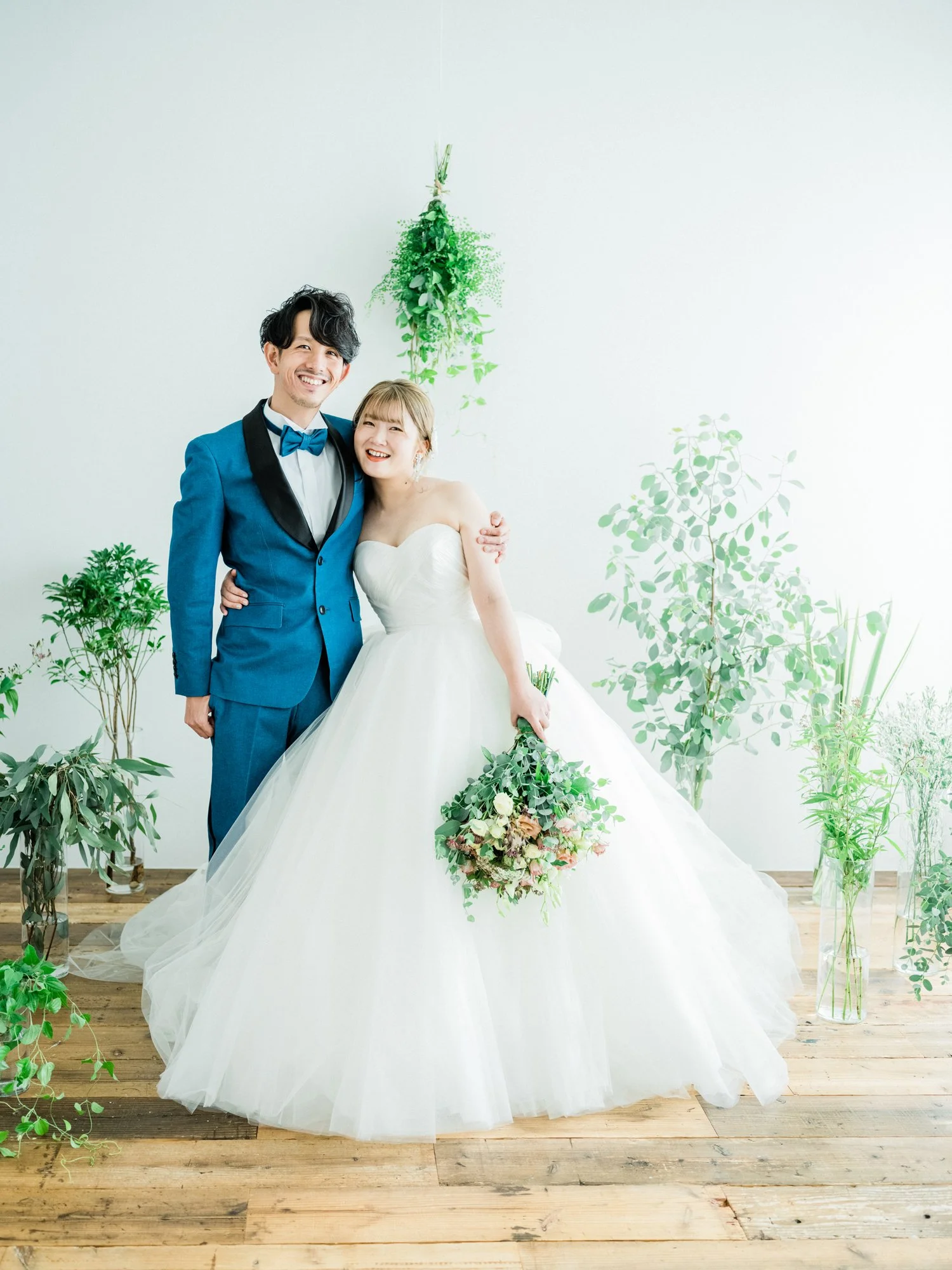 結婚式のカップルが笑顔で立っている写真。女性は白いウェディングドレスを着て、花束を持っている。男性は青いタキシードを着ている。背景には緑の植物が飾られている。