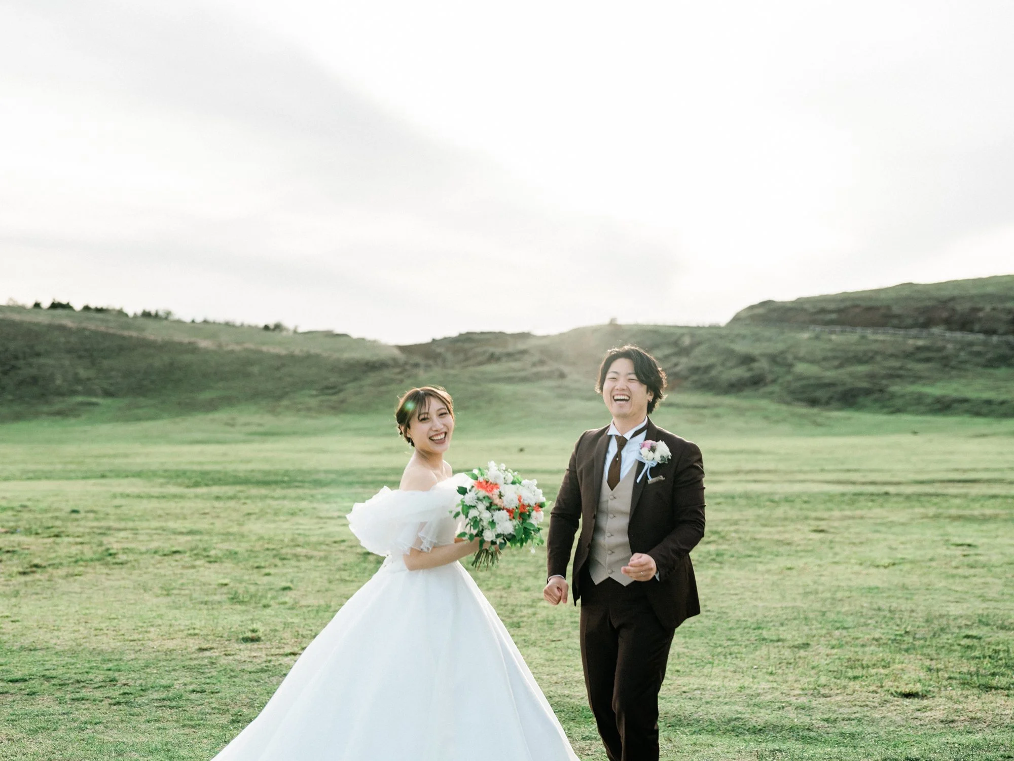 結婚式を挙げている新郎新婦が緑の丘の上で笑顔で手を取り合っている写真。