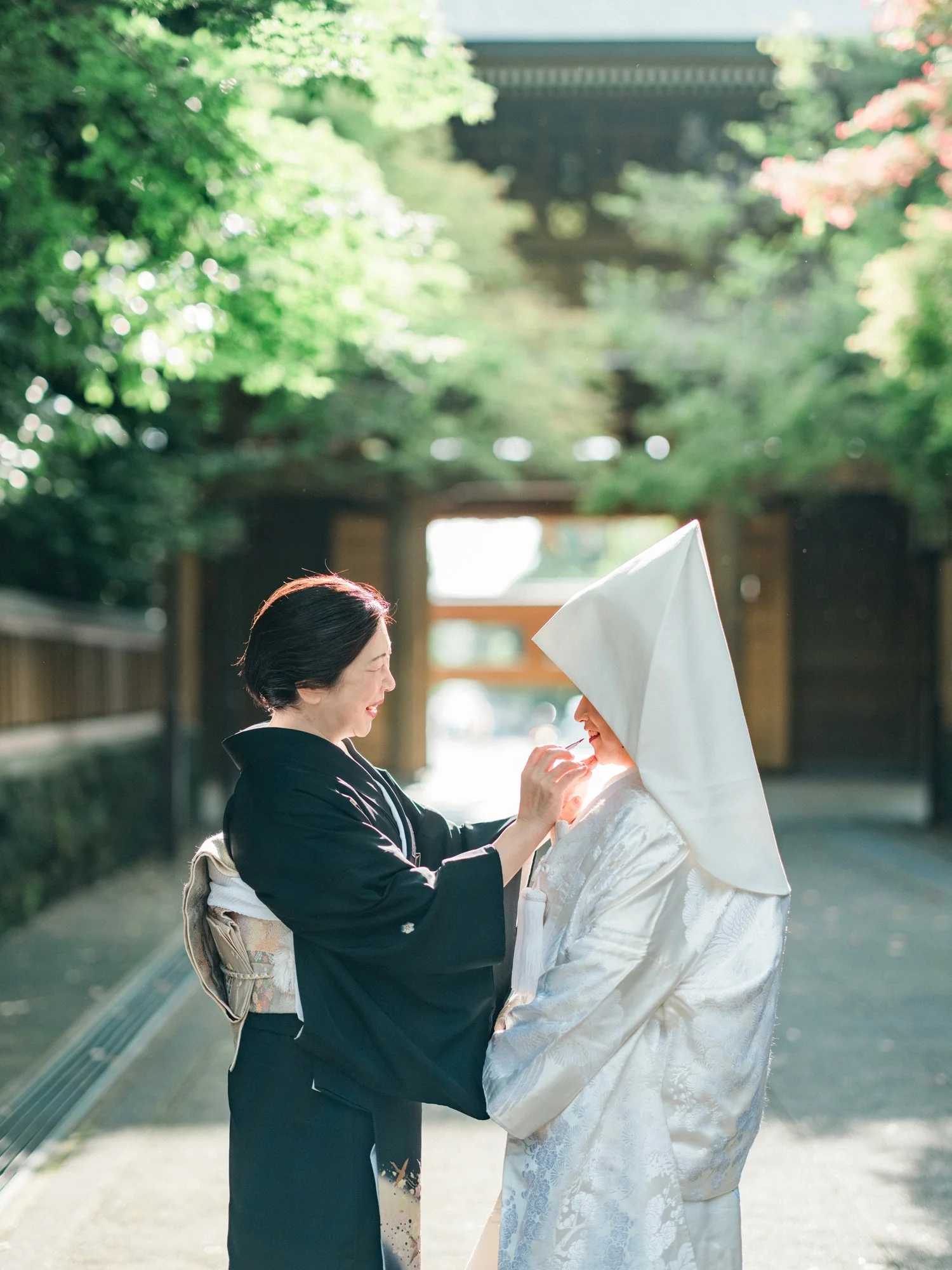 和装の花嫁と黒留袖のお母様が笑顔で交流しているシーン。伝統的な神社の境内で行われている。