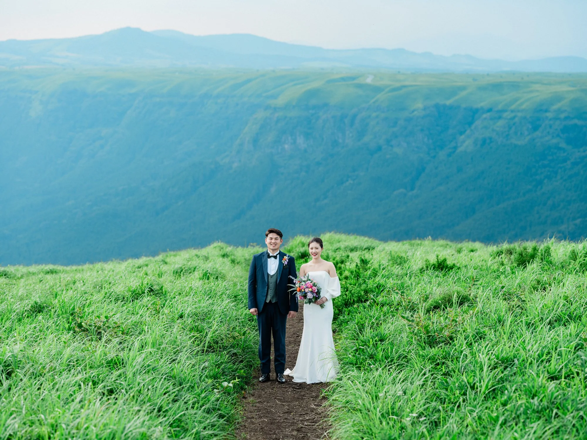 結婚式を挙げた新郎新婦が緑の草原を歩いている風景。背後には青い山々が広がる自然豊かな風景。